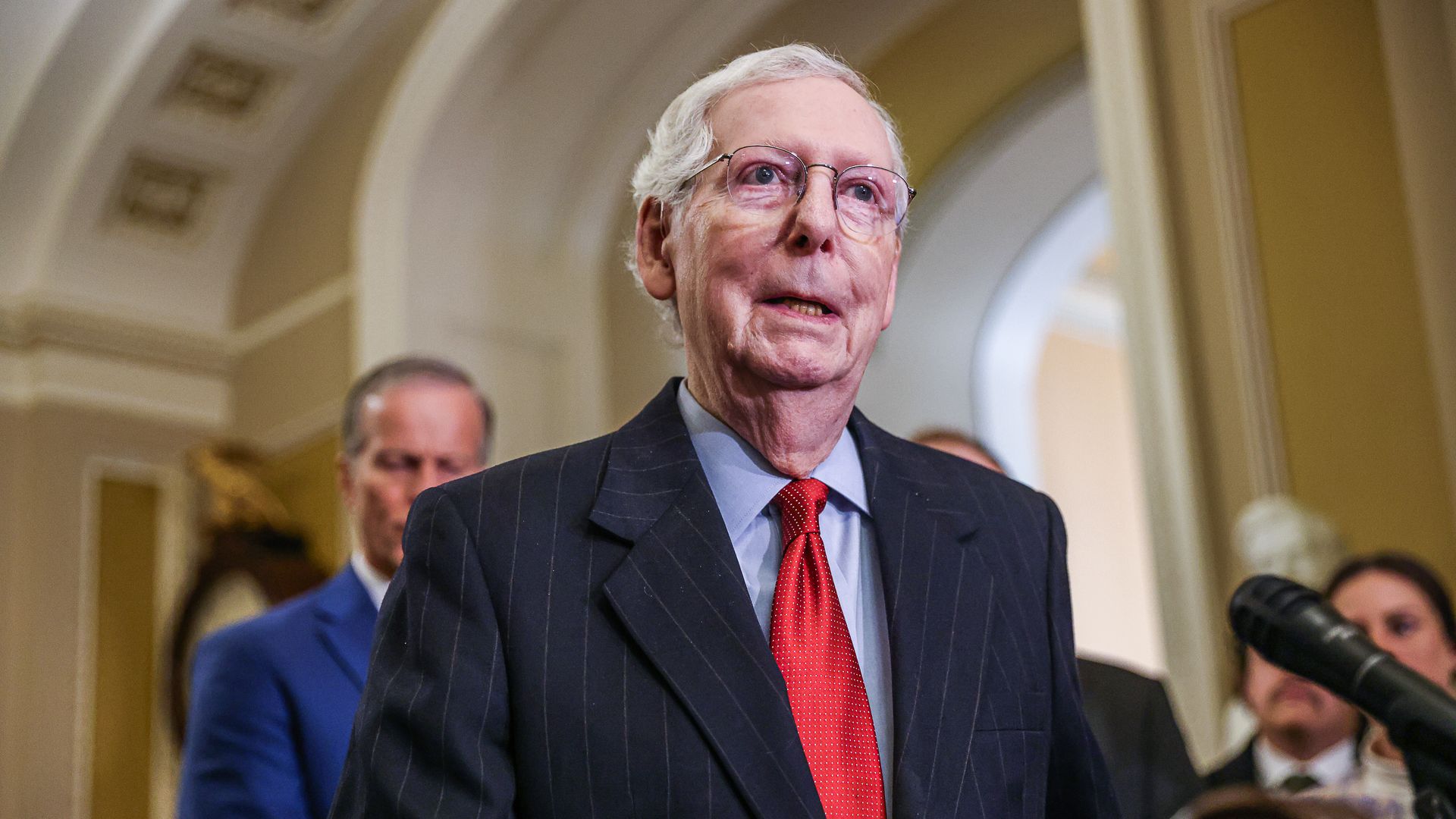 Mitch McConnell stands at a microphone wearing a dark suit, blue shirt and red tie. A few people are in the background. 