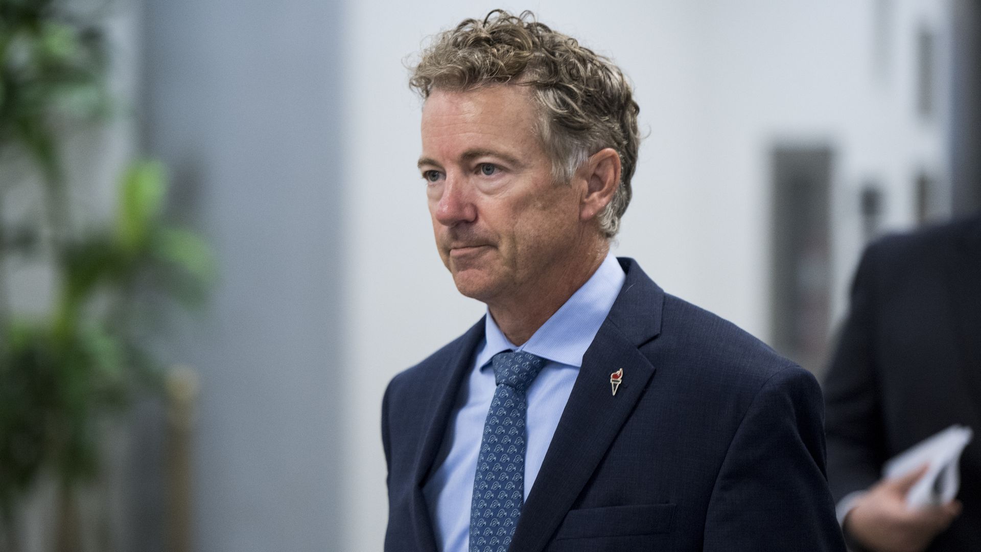 In this image, Rand Paul wears a suit and tie and walks down a hallway.