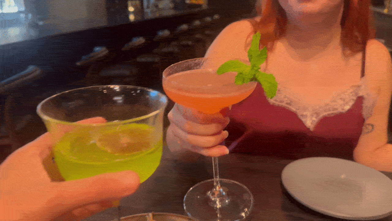 Two people toasting at a bar: a bright green lime cocktail in a tall glass on the left, and a pink cocktail with a mint sprig in a coupe glass held by a woman in a burgundy lace top.