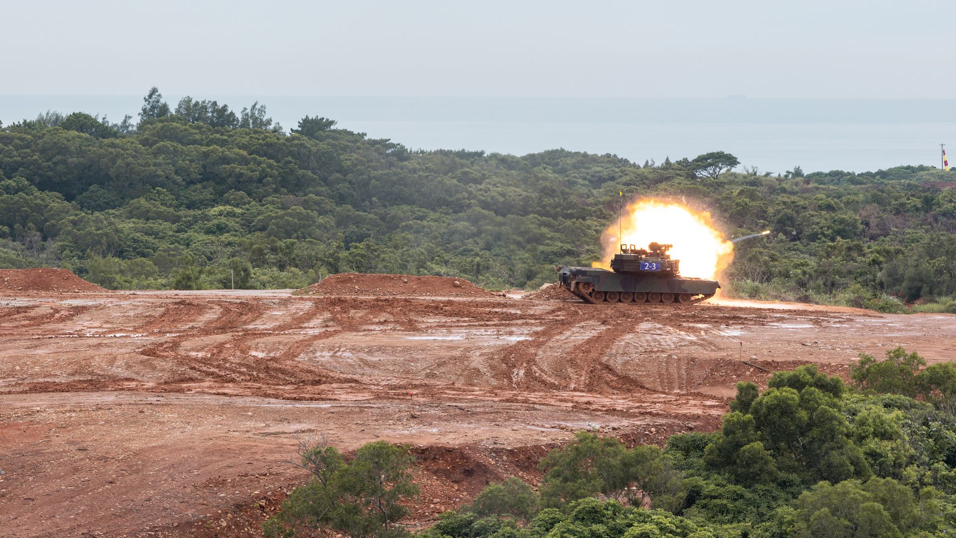 A tank fires its main gun after driving through muddy ground. A forest and shrubs is seen in the middle of the photo.