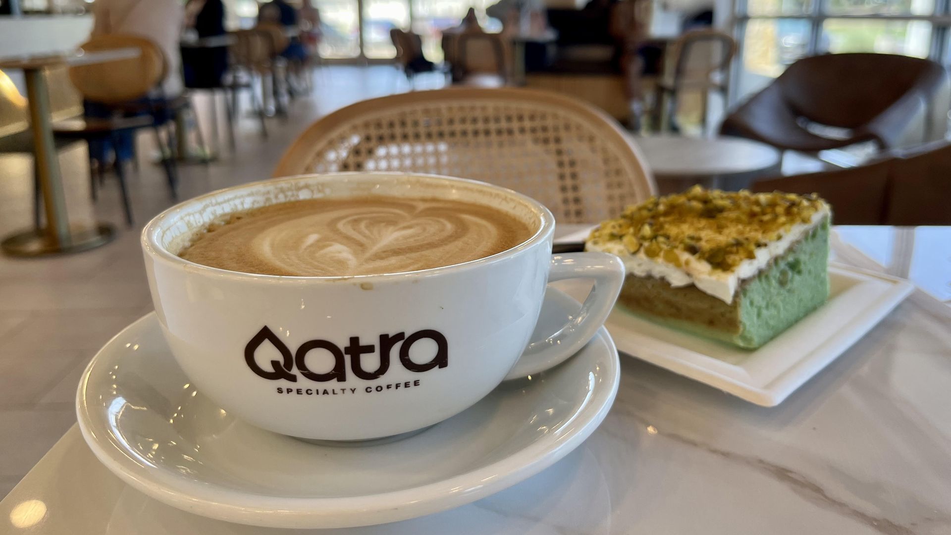 White coffee cup with latte art and labeled "Qatra Specialty Coffee" beside a slice of green cake with white frosting and crushed nuts.