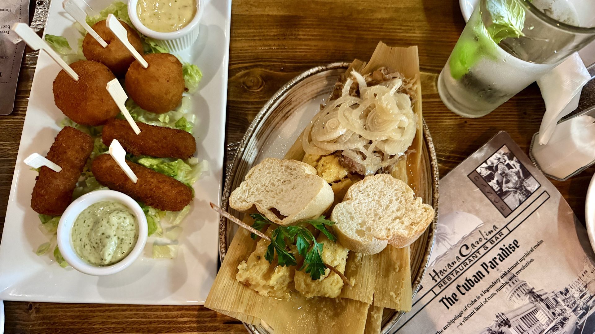Top-down view of a wooden table: left plate with croquettes on lettuce, sauces and skewers; right board with onion-topped flatbread and bread slices; a lime drink and vintage menu nearby.