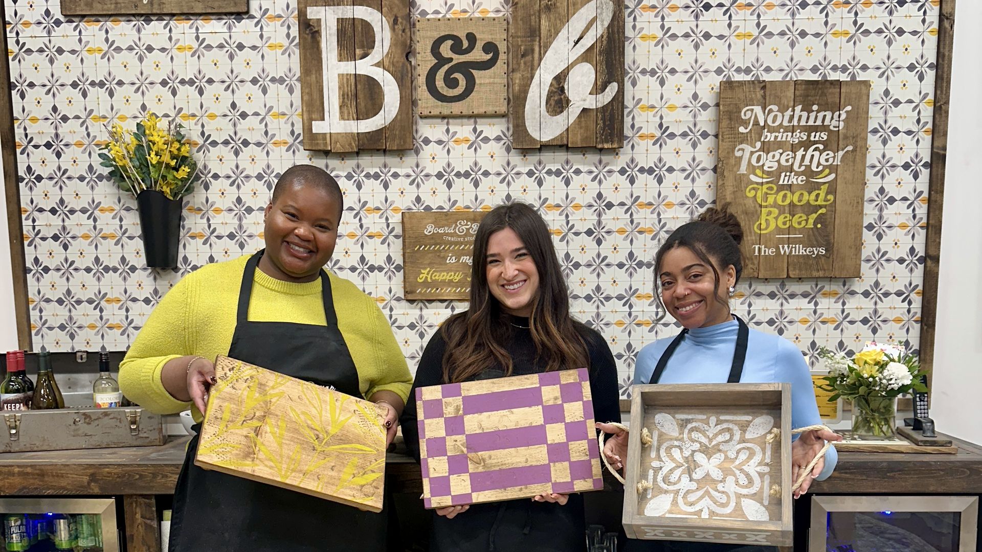 Board & Brush. Three smiling women in aprons pose behind a wooden bar, each holding a decorated board: yellow bamboo, purple-and-gold checker, and a white floral tile. A tiled wall with rustic signs behind them.