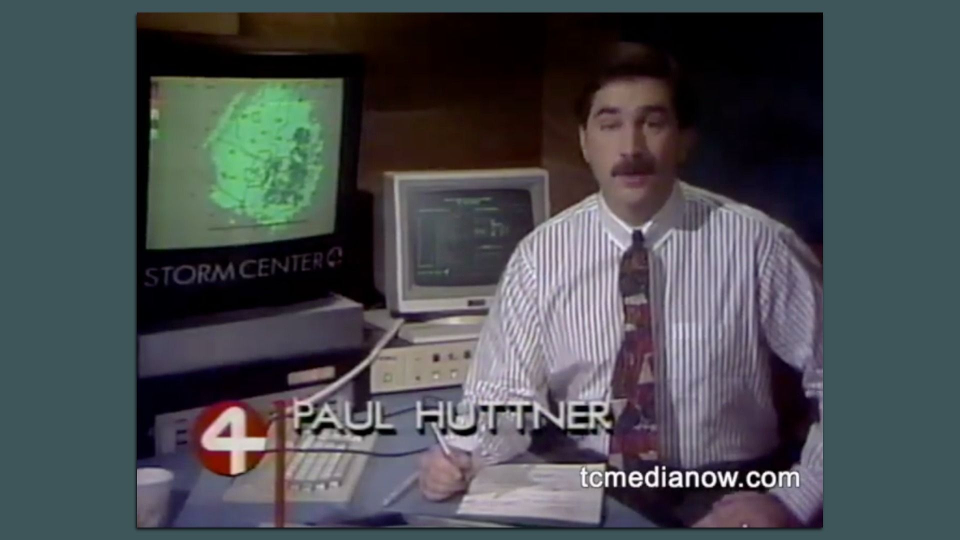 Man with a mustache in a white striped shirt and colorful tie sits at a desk with a pen and paper. Behind him are two monitors, one displaying a green weather map labeled STORM CENTER.