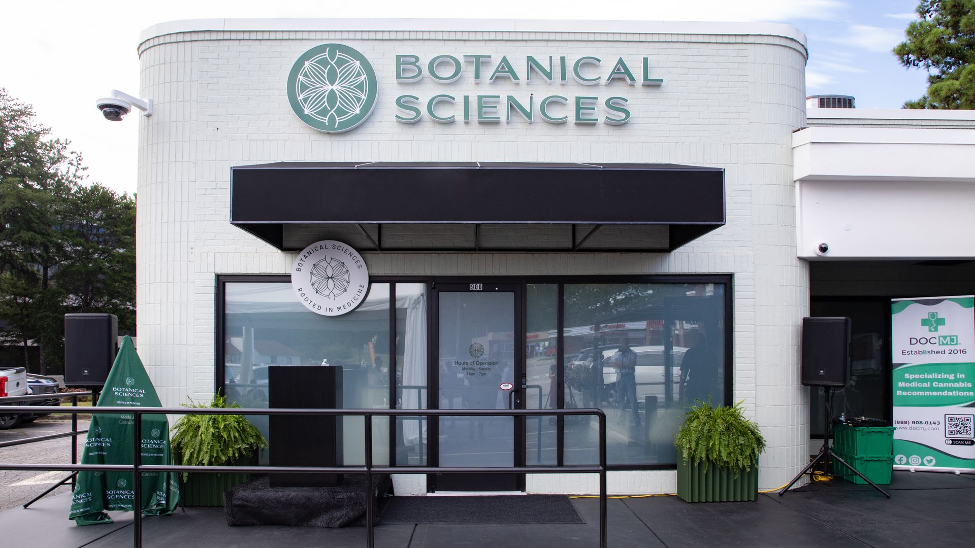 White brick storefront with a teal circular logo and teal text reading BOTANICAL SCIENCES; black awning over glass doors, a security camera on the left, and a blue sky with trees in the background.