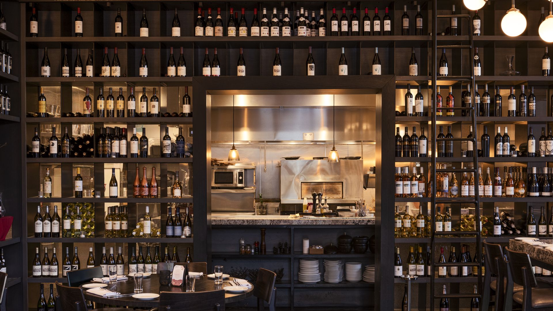 Cozy restaurant interior with shelves filled with wine bottles, a window into a stainless steel kitchen, and tables set with plates, glasses, and silverware in warm lighting.