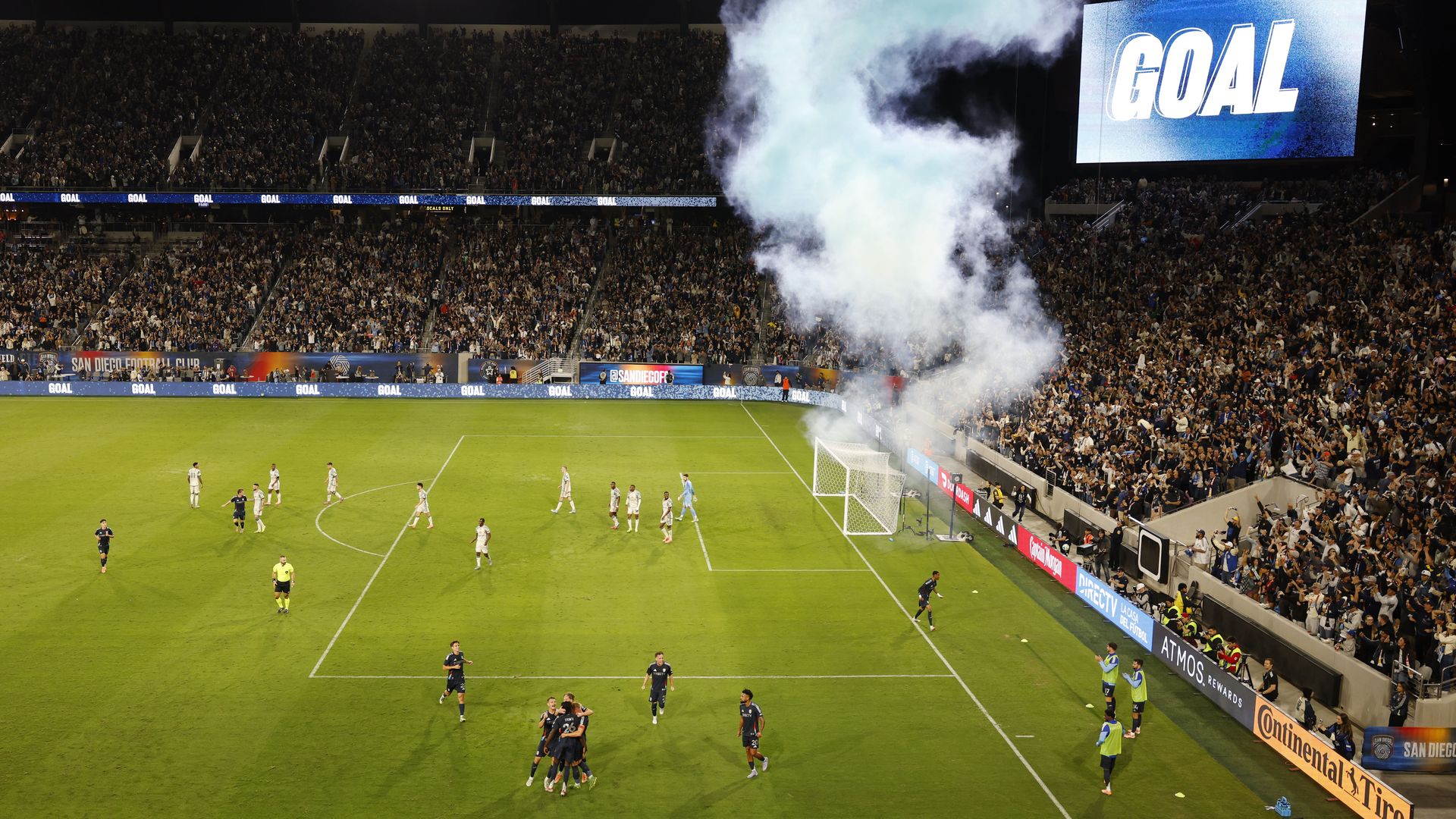 Soccer players celebrate on the field after scoring a goal as blue smoke rises behind the goal and fans cheer in the full stands of Snapdragon Stadium. 