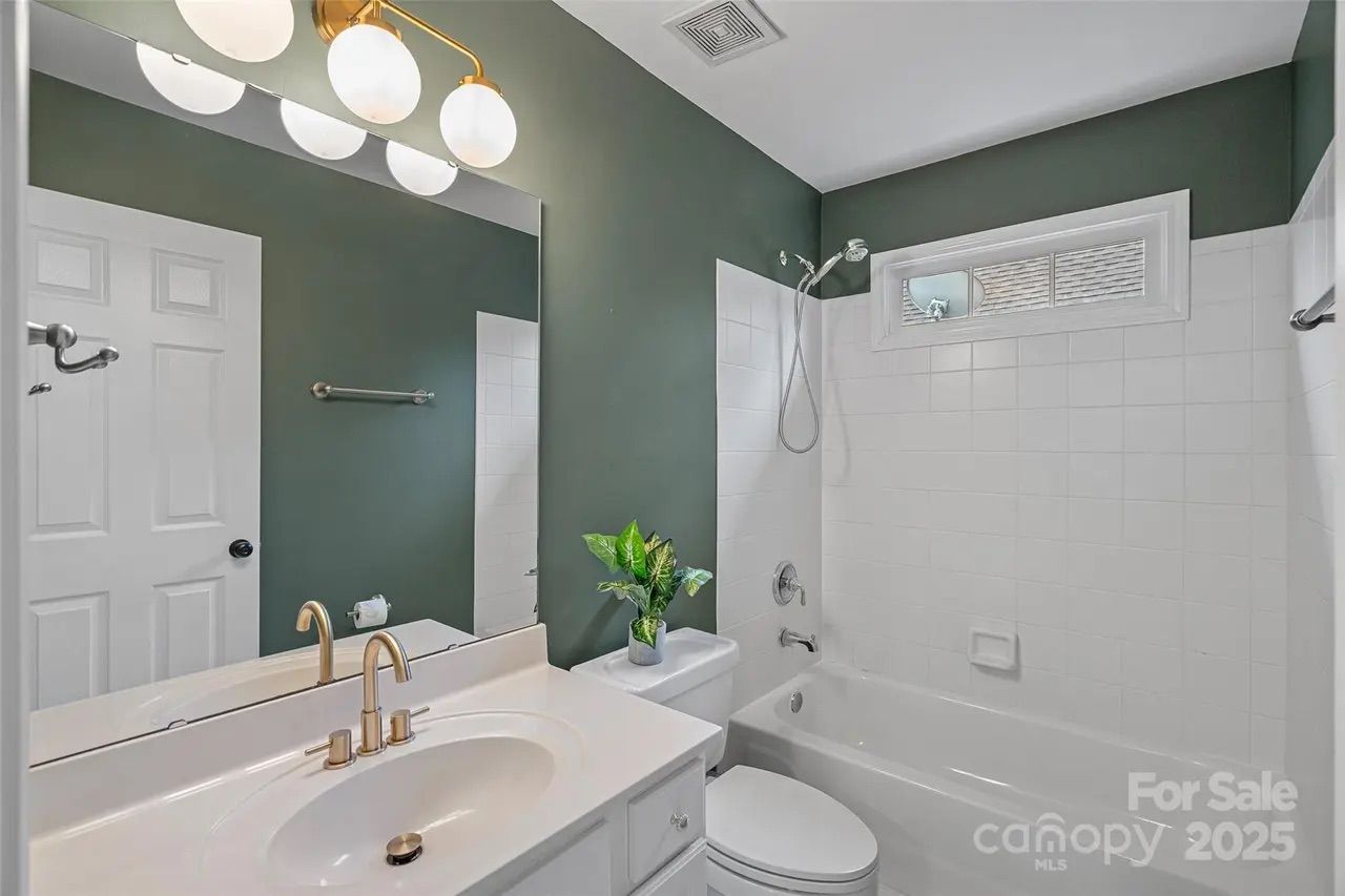 Bathroom with white bathtub and tiles, green walls, white vanity with gold faucet, round lights above mirror, white door, and a potted plant on the toilet tank.
