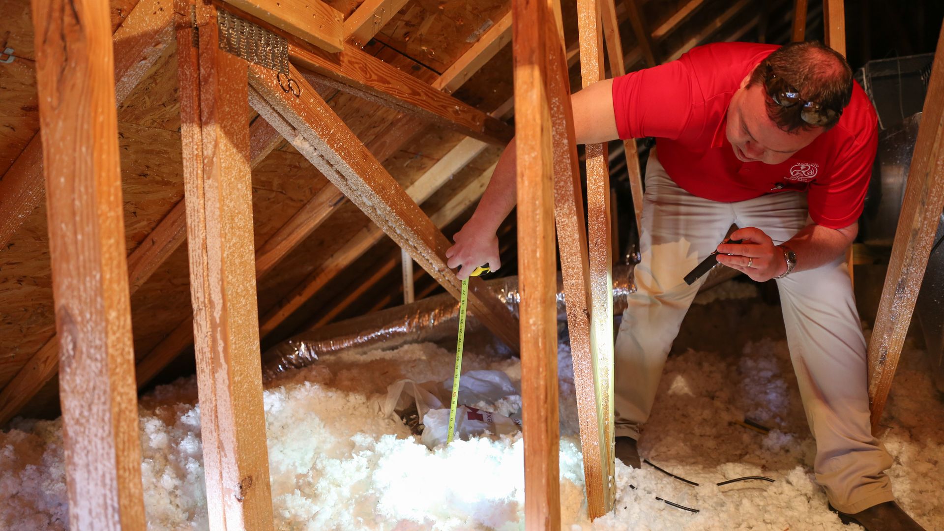 Man in a red polo shirt crouches in an attic among exposed wooden studs, wearing safety goggles and using a tape measure over scattered insulation.