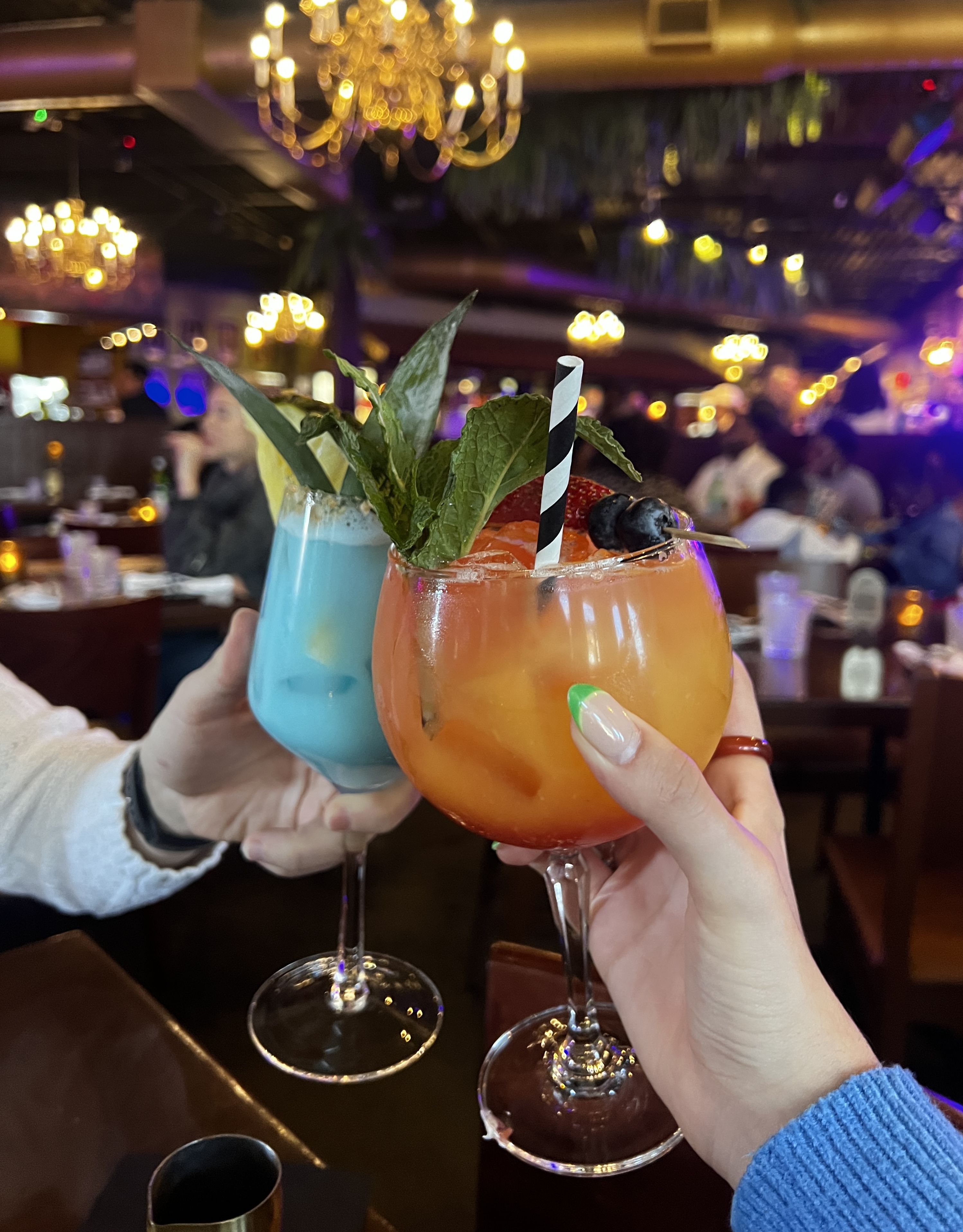 Two people clinking glasses in a lively restaurant setting. One glass contains a blue cocktail garnished with green leaves, while the other holds an orange drink with a black and white straw and a fruit skewer on top. Elegant chandeliers and warm lighting enhance the atmosphere in the background.