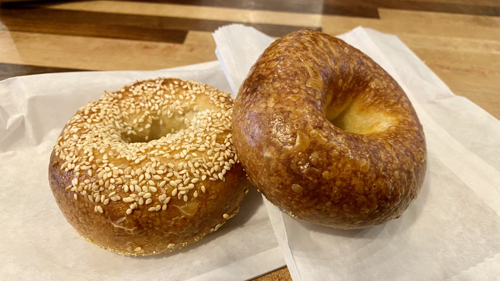 Toasty looking bagels, one plain, one with sesame, on a paper sleeves atop a wooden table.