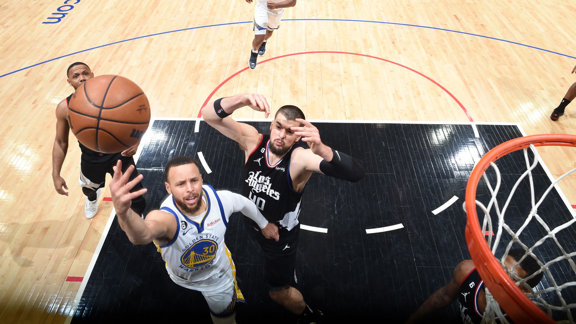 A shot from above showing Steph Curry, left, shooting a basketball into the basket on the right. One LA Lakers player jumps up trying to stop him. Another on the right watches the shot happen.