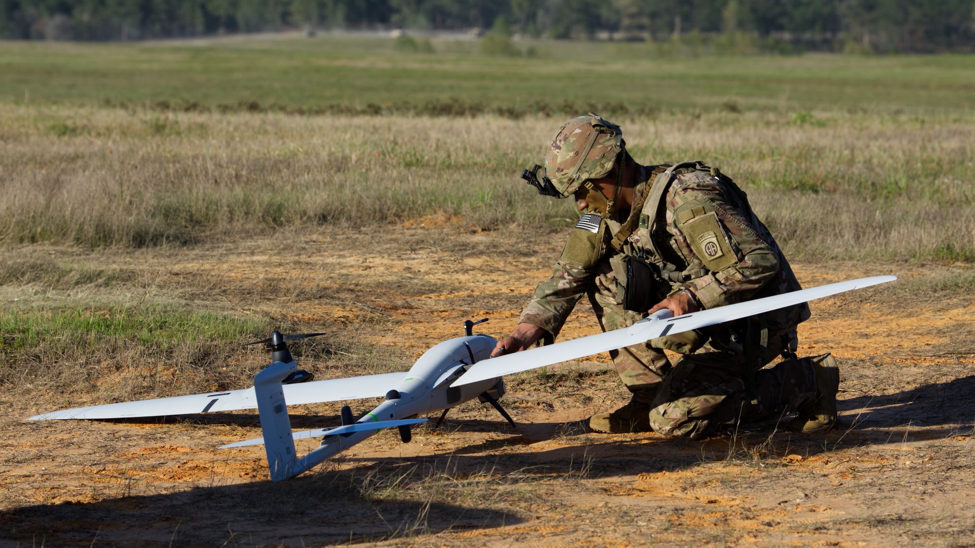 Camouflage-suited soldier kneels in a dry field, inspecting a white fixed-wing drone with long wings. He wears a helmet and tactical gear, with an American flag patch visible on his sleeve.