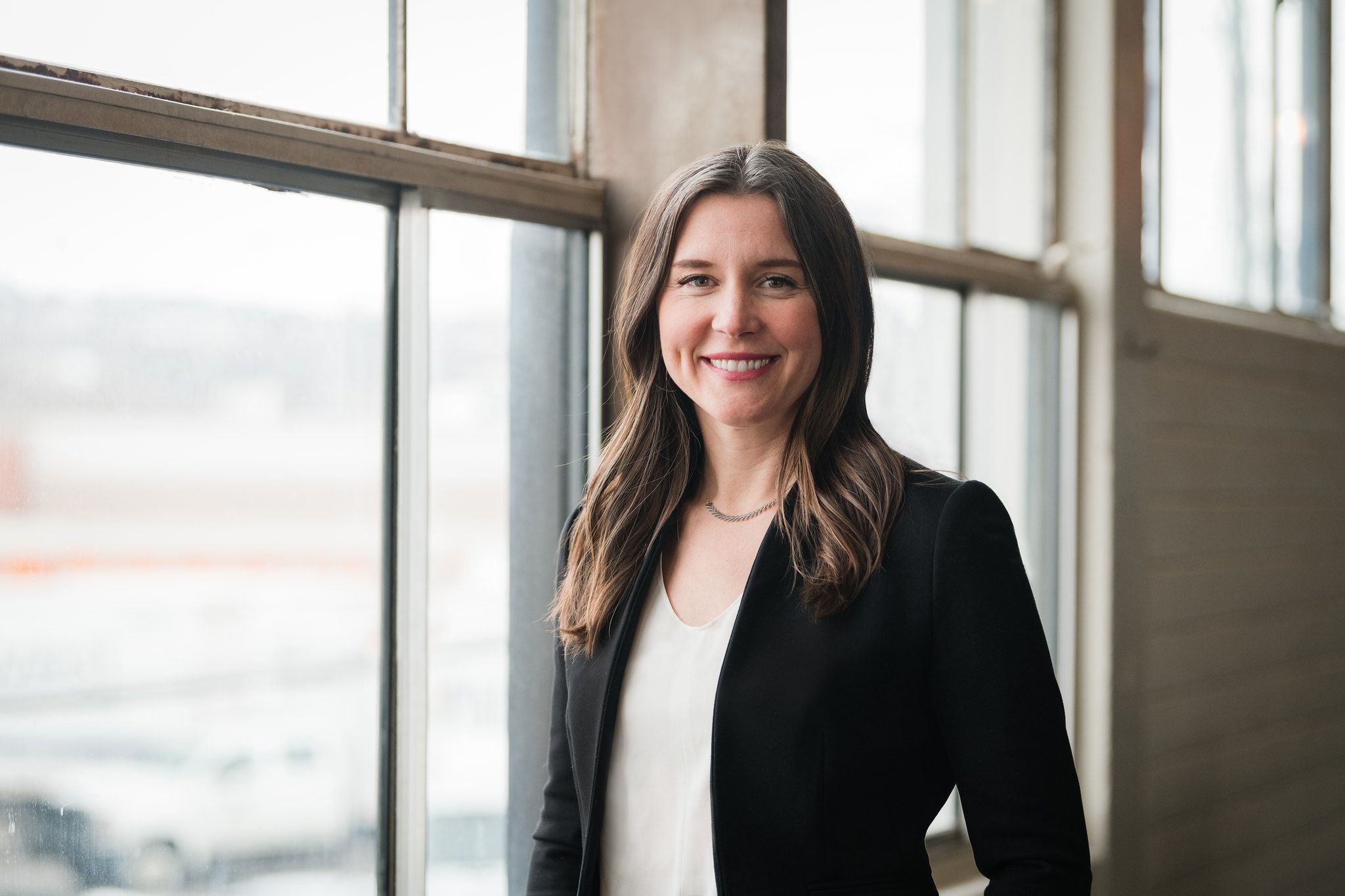 A photo of a woman with a black blazer and white shirt next to windows.