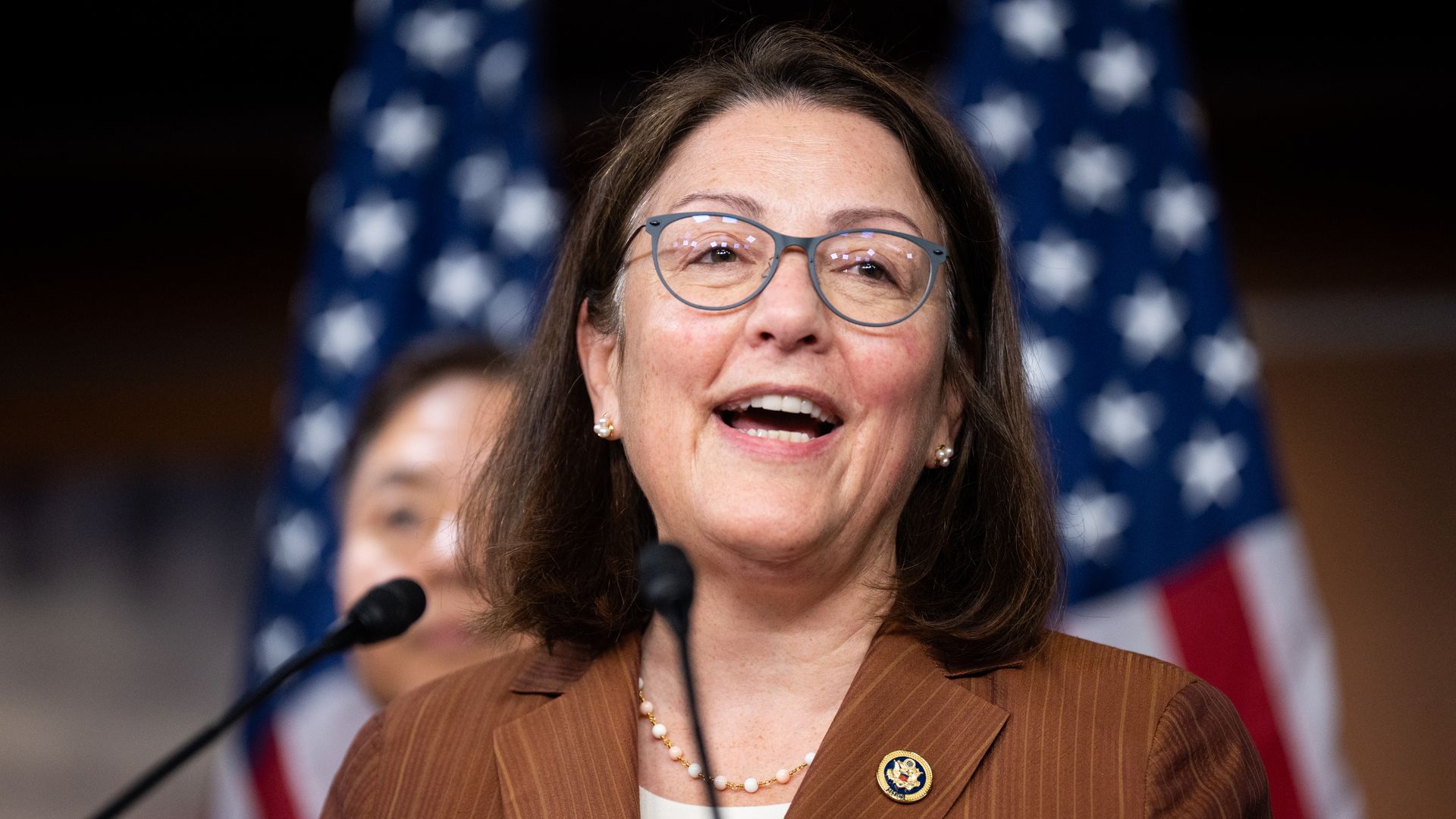 Rep. Suzan DelBene, wearing a dark orange blazer and standing in front of American flags.