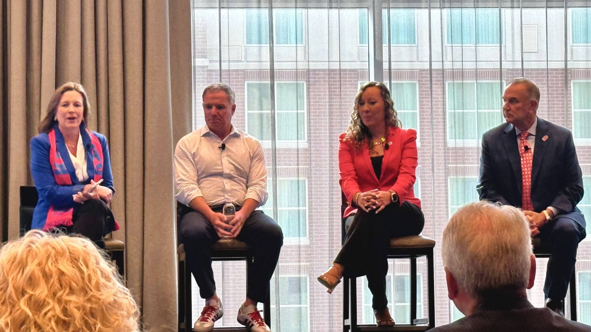Panel of four on stools in front of tall windows; left to right: woman in blue blazer with pink scarf, man in white shirt, woman in red blazer, man in dark suit with pink tie; audience in foreground.