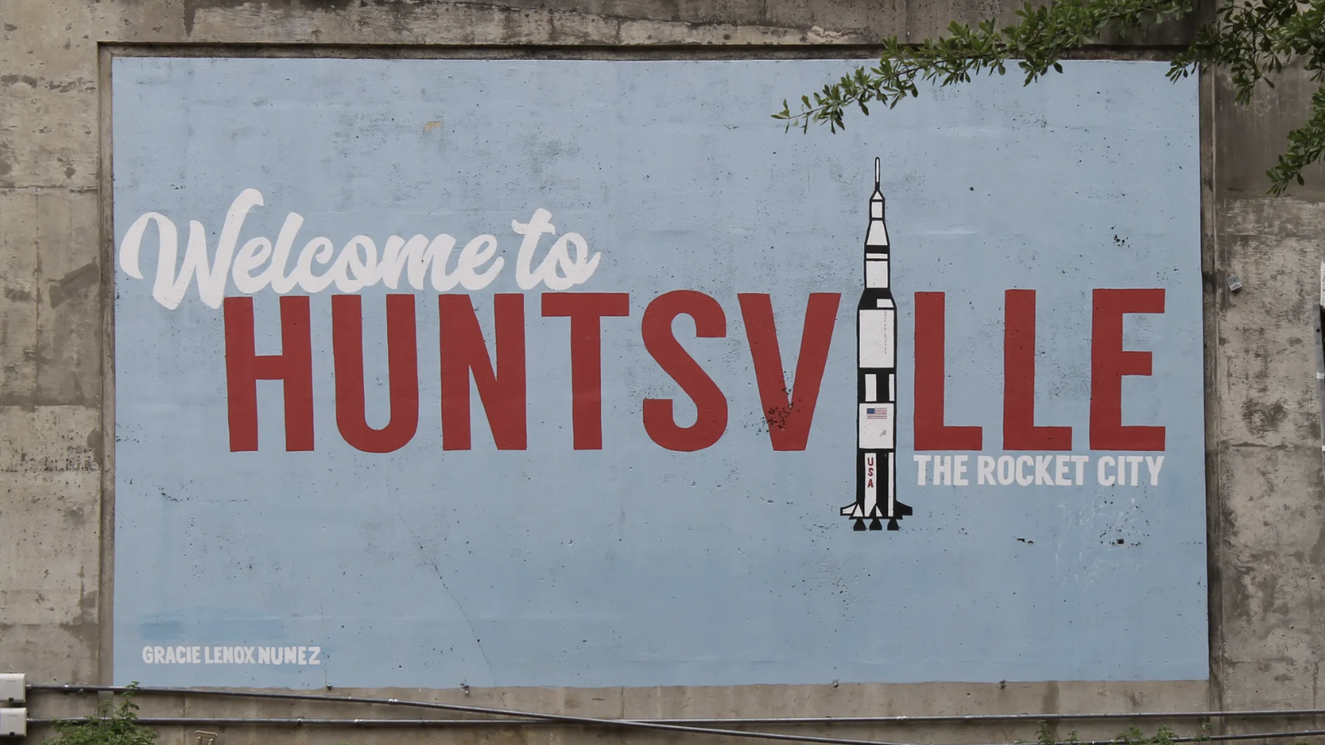Mural on concrete wall reading "Welcome to Huntsville" in red and white letters with a rocket illustration and "The Rocket City" tagline on a light blue background.