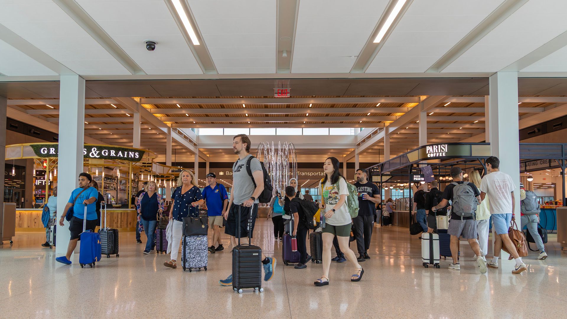 Busy airport terminal with travelers walking and pulling suitcases near signs for "Grains & Grains" and "Parisi Coffee" shops under bright ceiling lights.