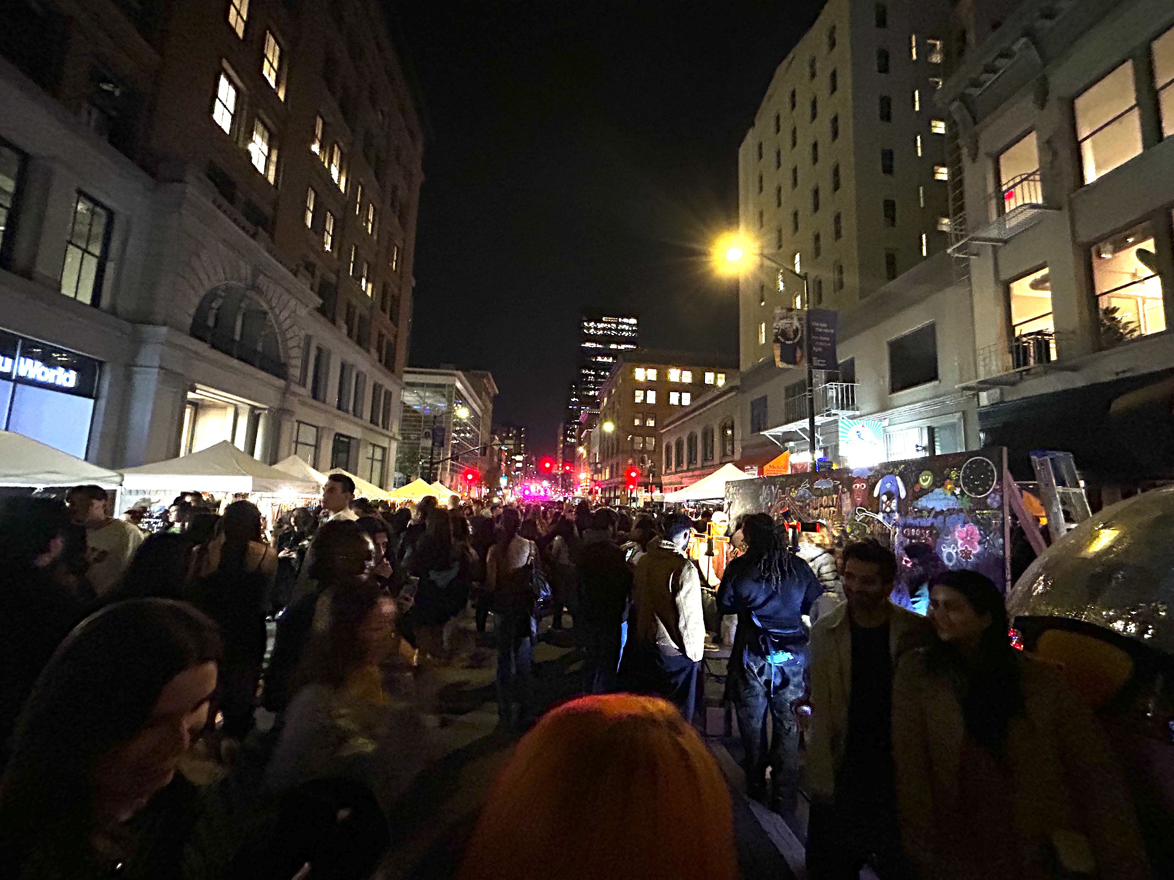 Nighttime street fair with many people walking between tents and illuminated buildings, colorful mural on the right, and city lights in the distance.