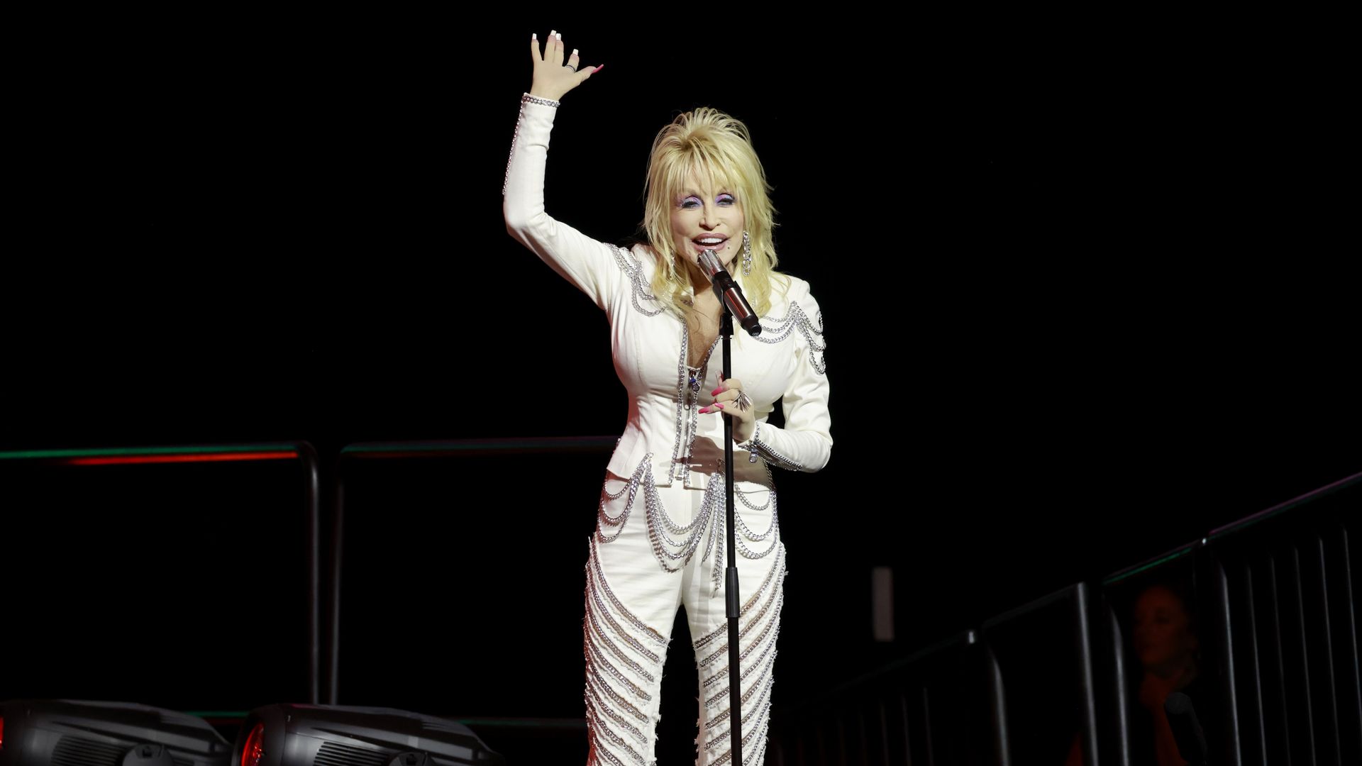 An image of Dolly Partin in a white suit decorated with chains waving to the audience from a microphone.