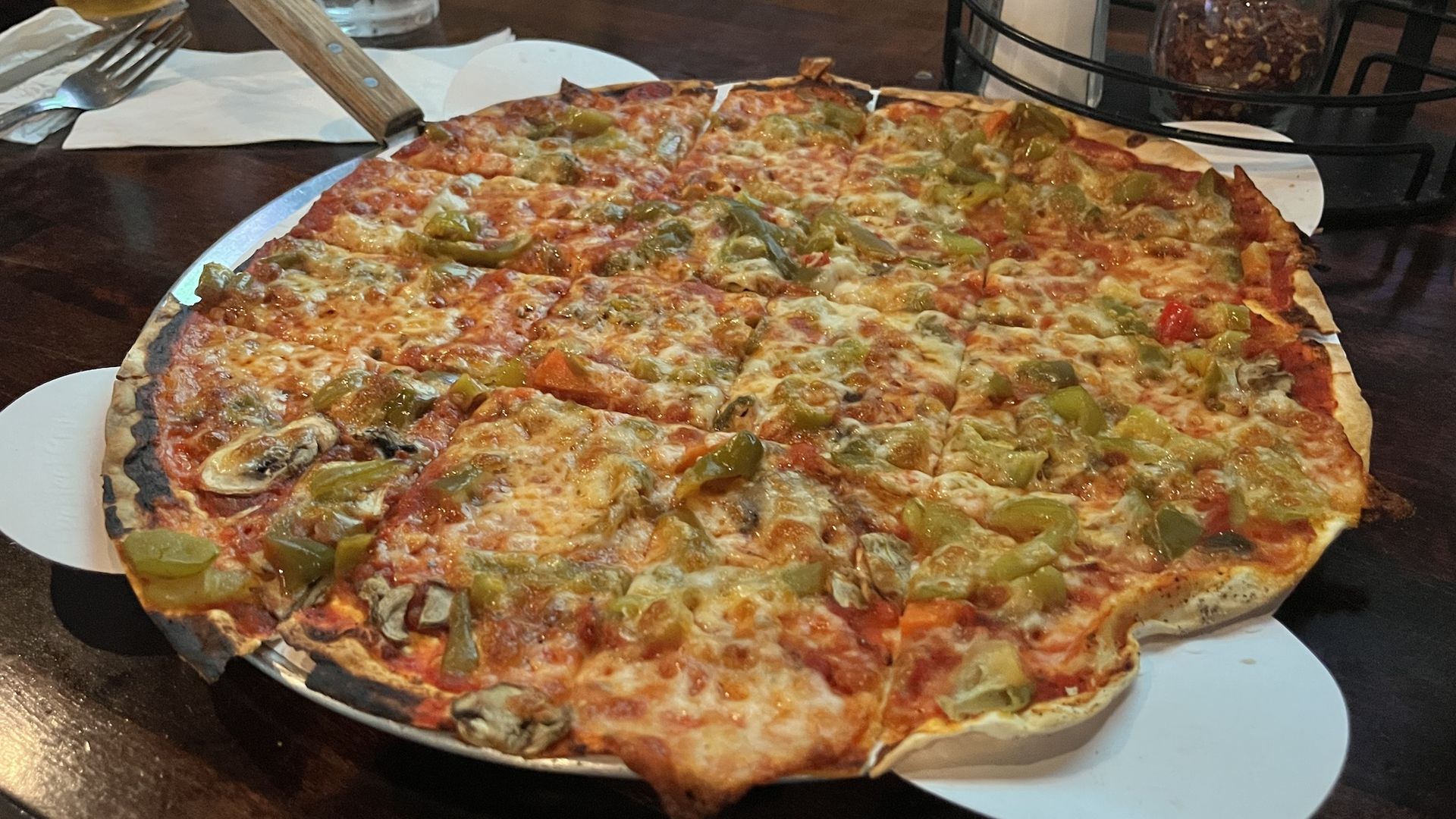 Large veggie pizza with green peppers and mushrooms, melted cheese, and a thin crust on a white plate. A wooden pizza server rests on the edge; a beer glass and condiment jars sit in the background.