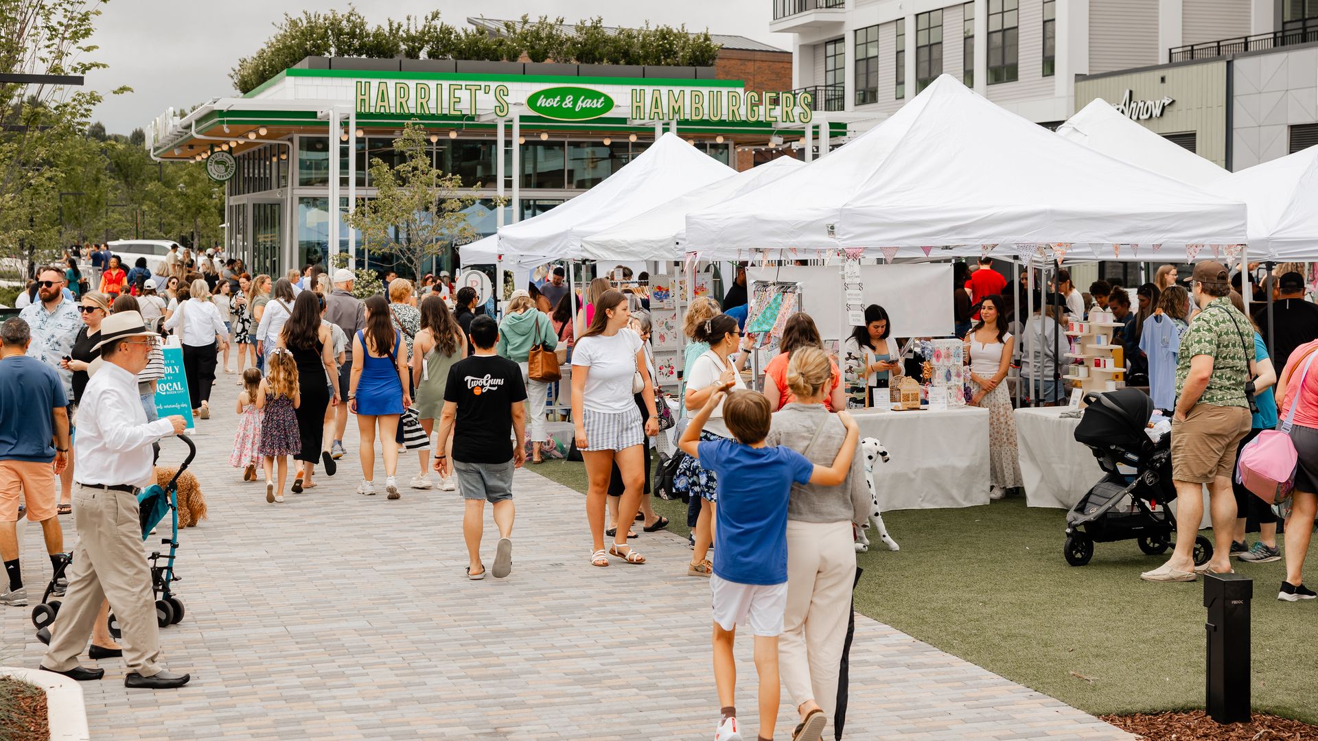Crowded outdoor market with white tents and people browsing, walking, and socializing near Harriet's Hamburgers restaurant under cloudy sky in an urban area with modern buildings.