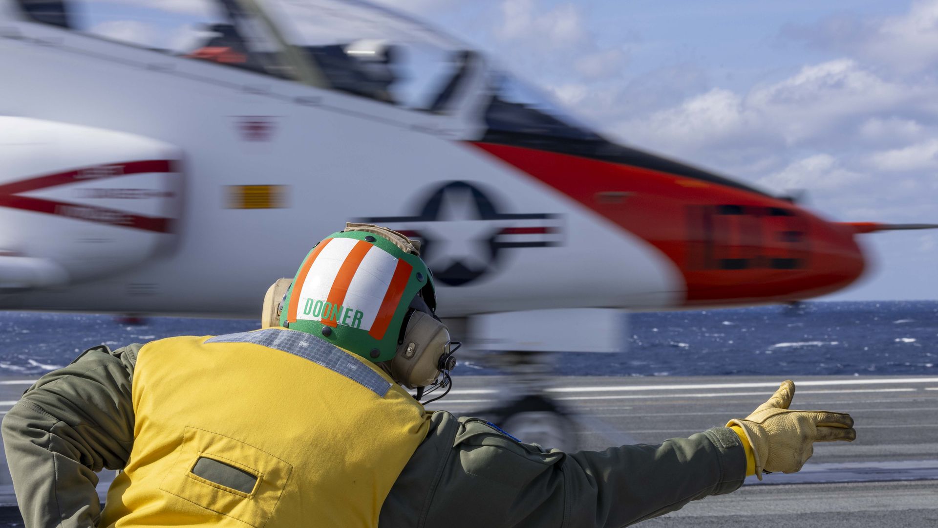 A man in a bright yellow vest and green helmet with orange stripes labeled DOONER extends an arm to guide a red-and-white aircraft on a carrier.