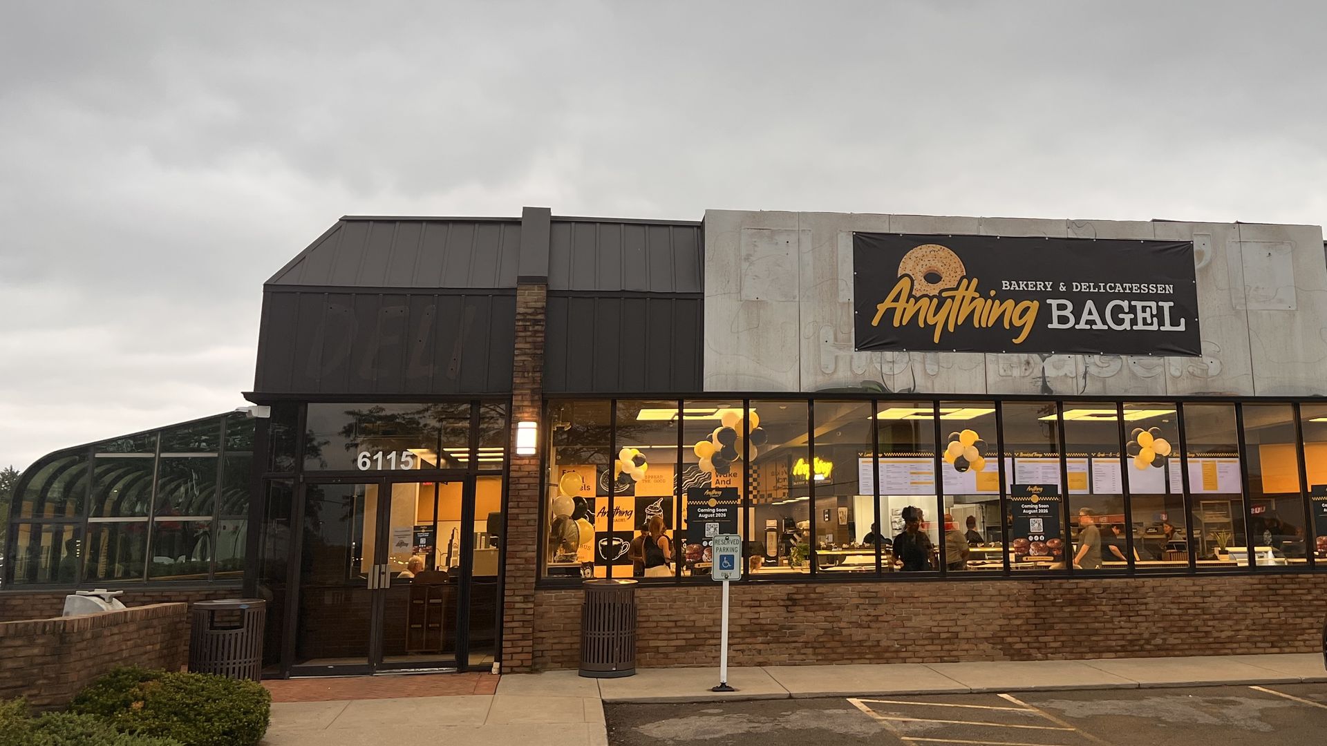 Overcast sky above a bakery storefront named "Anything Bagel" with an orange bagel logo. Glass windows show balloons inside; a parking lot and a gray car sit in front.