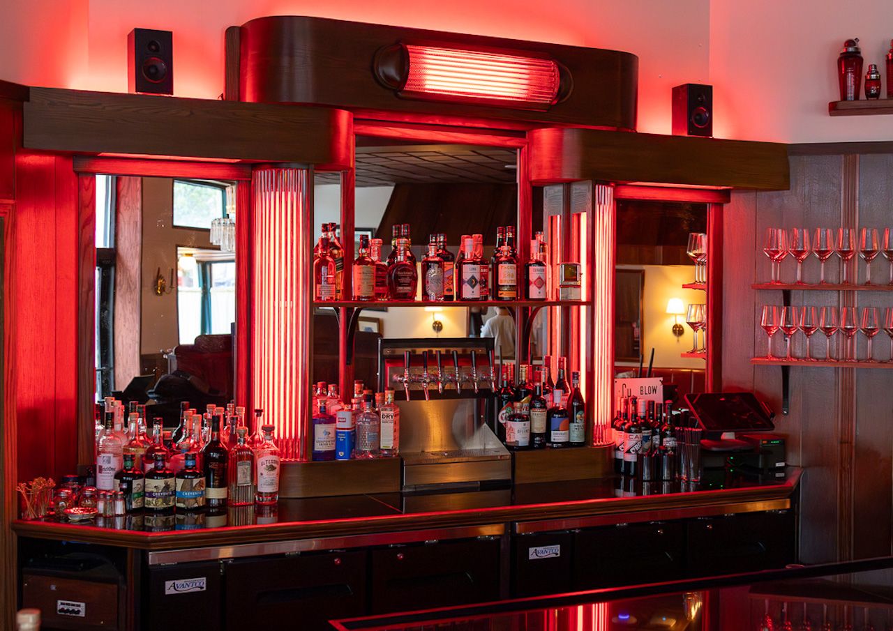 Bar interior bathed in red neon light; bottles line shelves behind a glossy counter, glassware on the right, and a large red-lit fixture above the back bar.