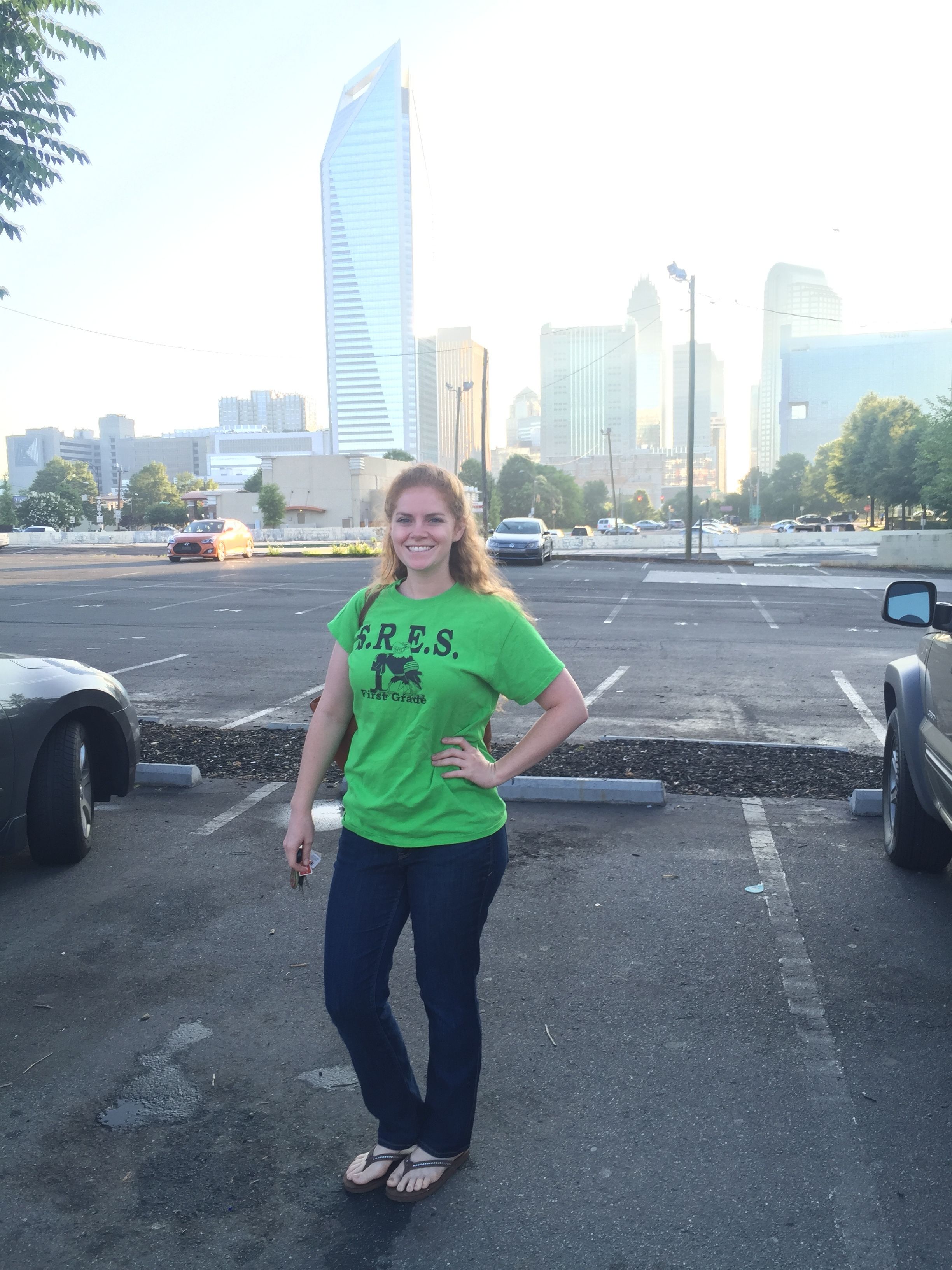 Woman in a bright green S.R.E.S. first grade T-shirt, jeans, and sandals smiling, standing in a parking lot with city skyscrapers and trees in the background during daylight.