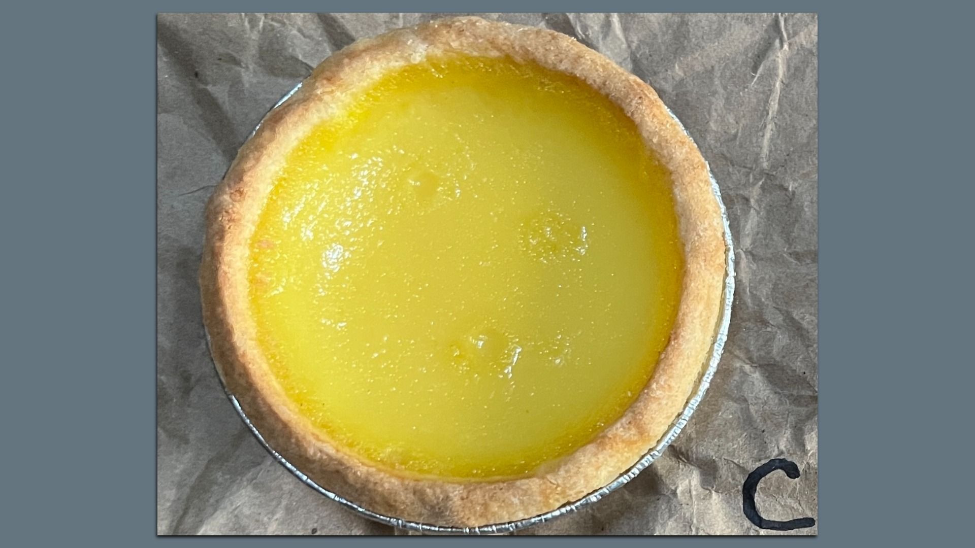 Top view of a small lemon tart with a bright yellow filling and golden brown crust in a foil container, placed on crumpled brown paper with a partial black letter visible.