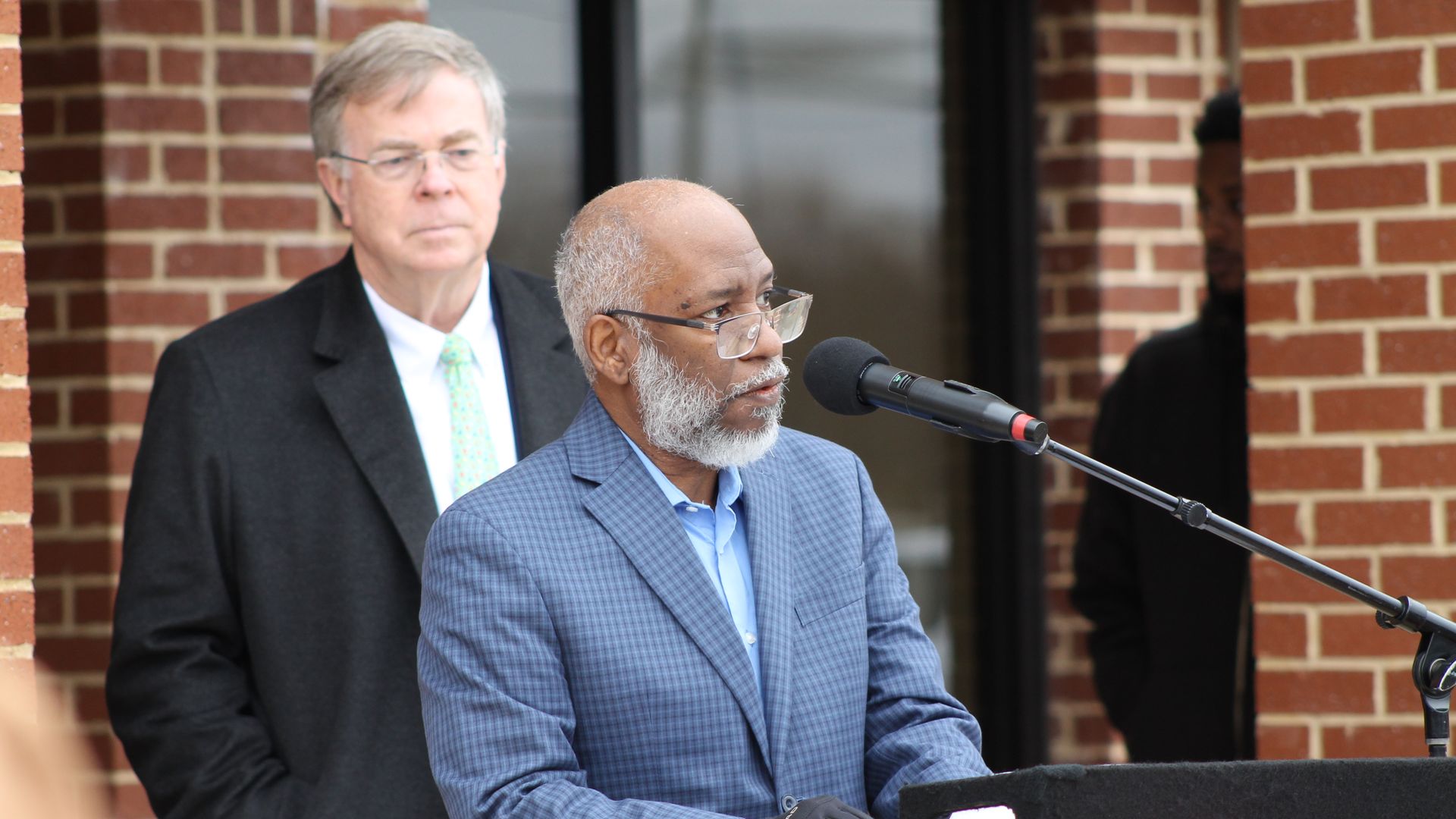An older man with glasses, a beard, and a blue checkered suit speaking at a microphone with a podium decorated with red ribbon, a man in a black coat stands behind him.