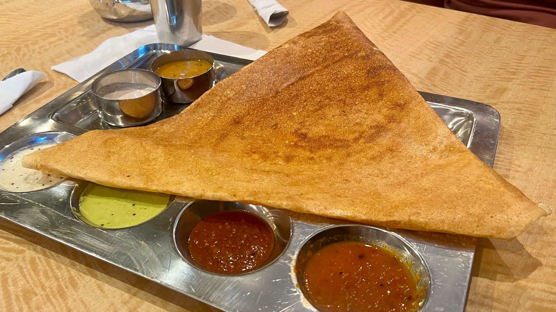 Triangular golden-brown dosa on a metal tray with five small bowls containing white, green, red, and brown chutneys and sambar on a wooden table with napkins and a steel cup in the background.