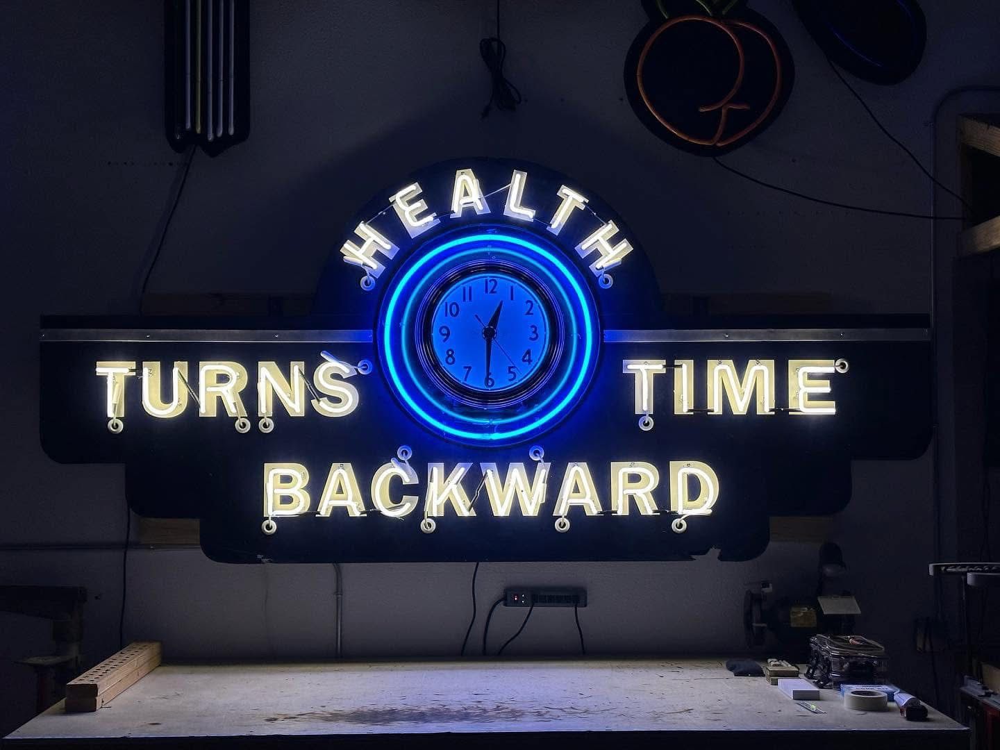 Neon sign reading "HEALTH TURNS TIME BACKWARD" with a bright blue circular clock at the center, mounted on a dark wall above a workbench in a workshop.