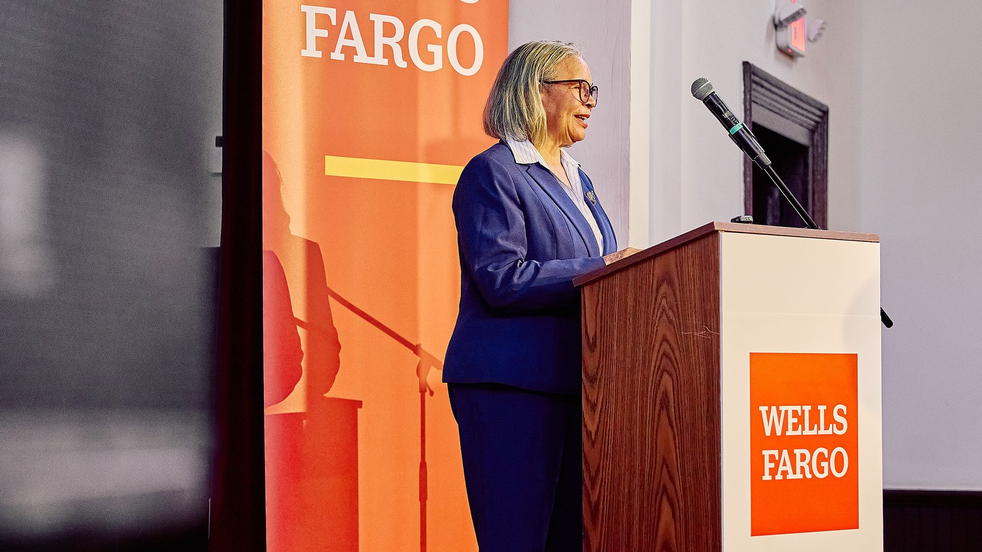A woman in a blue business suit and glasses speaks at a Wells Fargo event from a wooden podium with a microphone; an orange Wells Fargo banner is in the background.