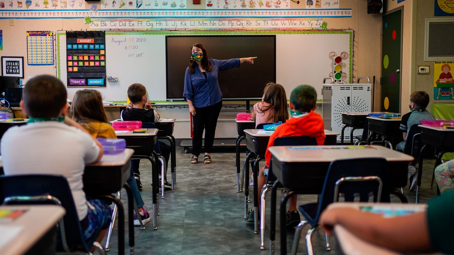 Photo of a teacher standing at the front of a classroom and pointing to their left as students sit at their desks in rows