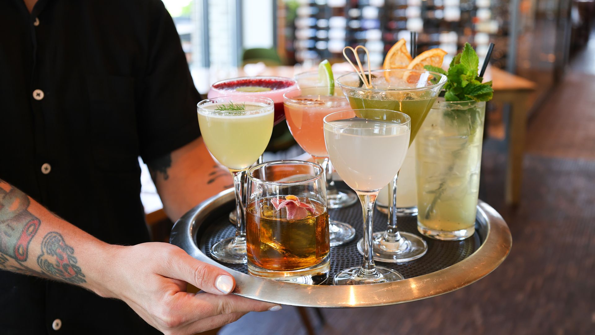 A person with tattoos and a black shirt holds a silver tray carrying eight colorful cocktails garnished with herbs, citrus, and flowers in different glassware inside a restaurant.