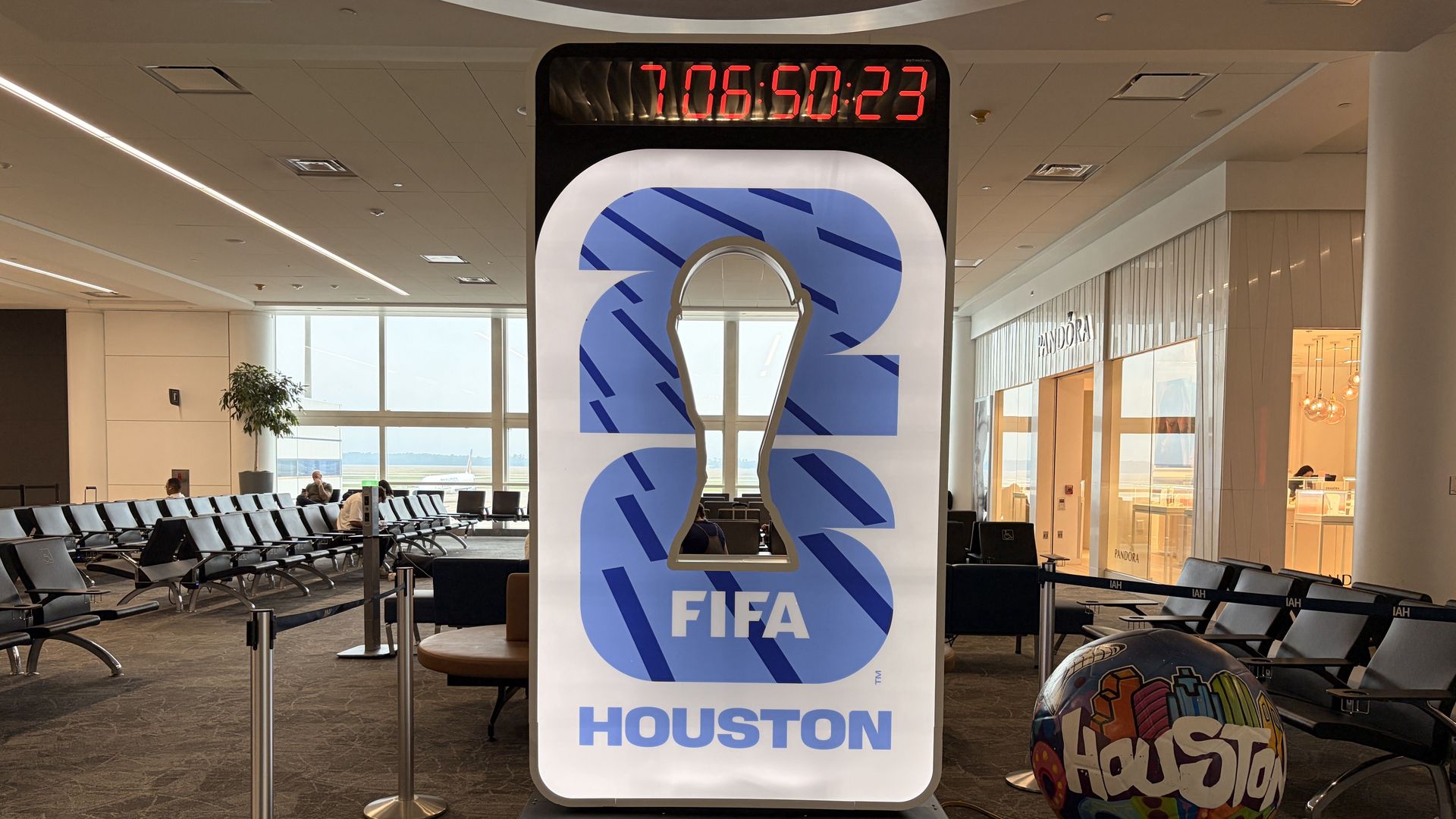 Inside an airport terminal, a tall FIFA World Cup 2026 display shows a blue question-mark design with a red countdown clock above. Seating, windows, Pandora store, and a colorful ball are in view.