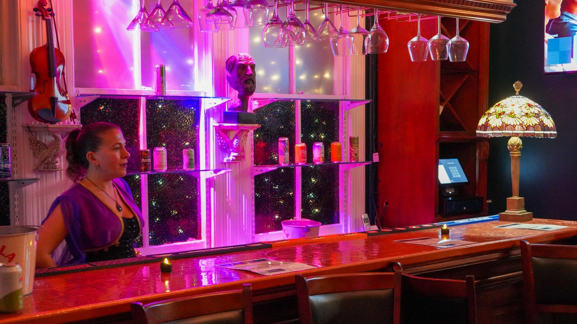 Woman behind a wooden bar with purple lighting, hanging wine glasses, a violin on wall, and a stained-glass lamp on the bar in a cozy room.