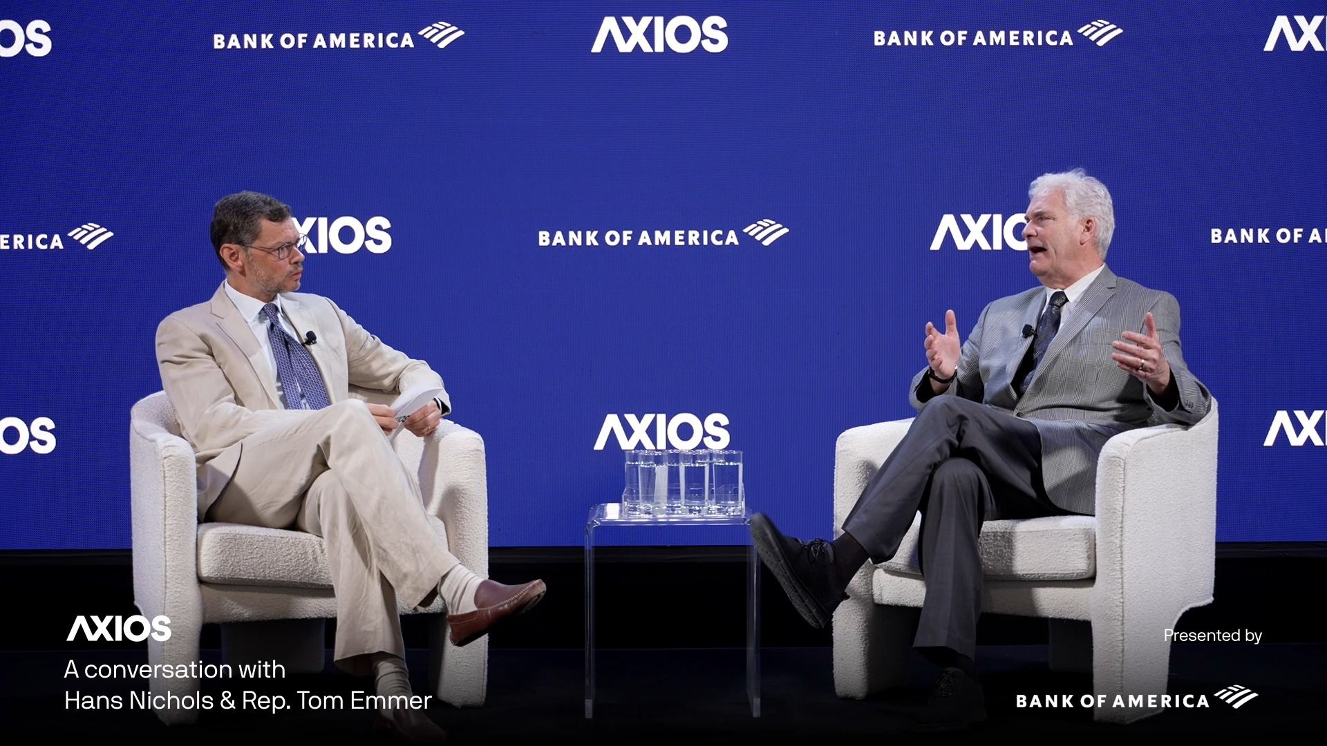 Two men in suits seated on white chairs on a stage with a blue backdrop displaying AXIOS and Bank of America logos, engaged in a conversation during an event.