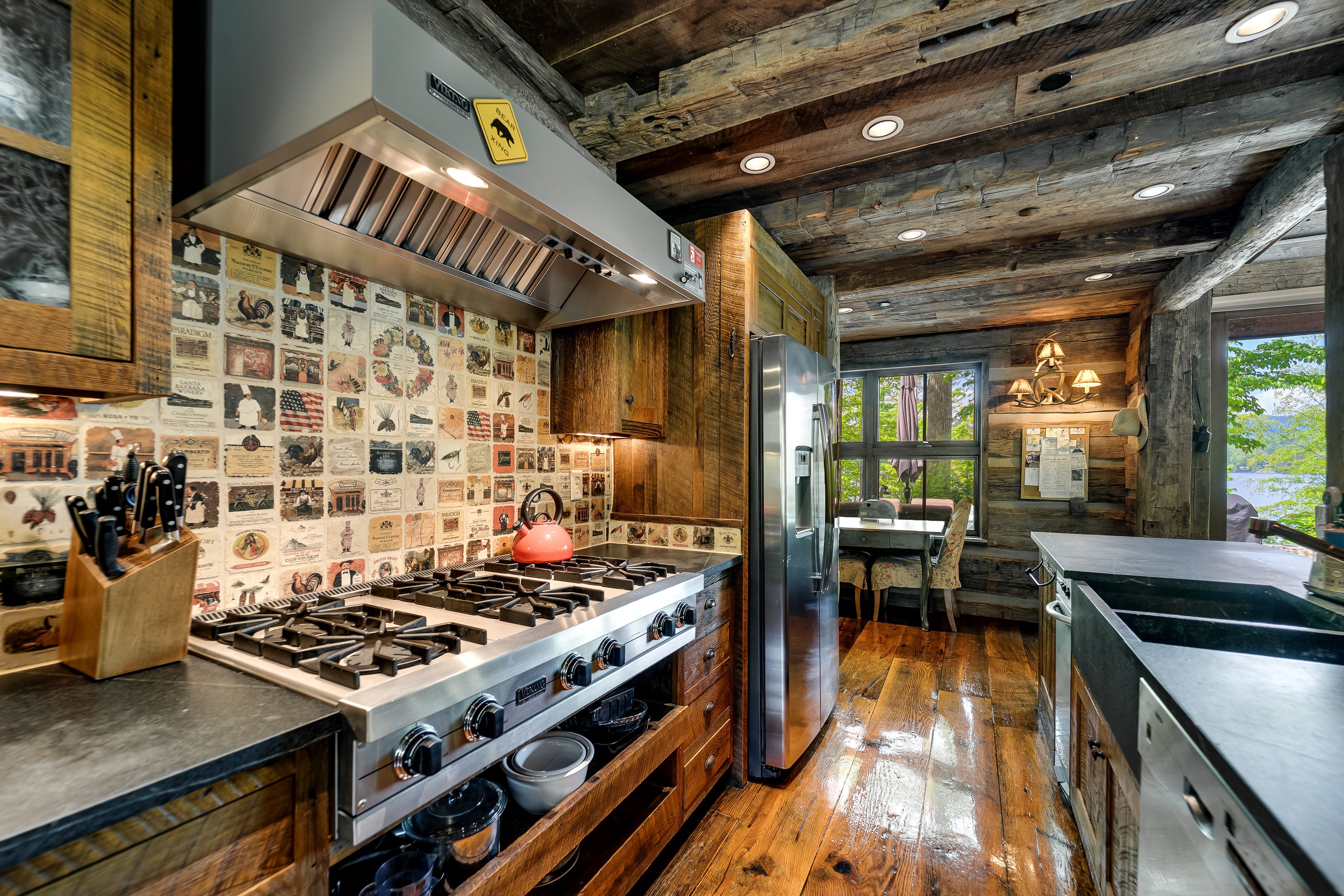 Rustic kitchen with wooden beams, hardwood floor, stainless steel stove and fridge, a red kettle on the stove, decorative tile backsplash, and a dining area by large windows with views of trees and water.