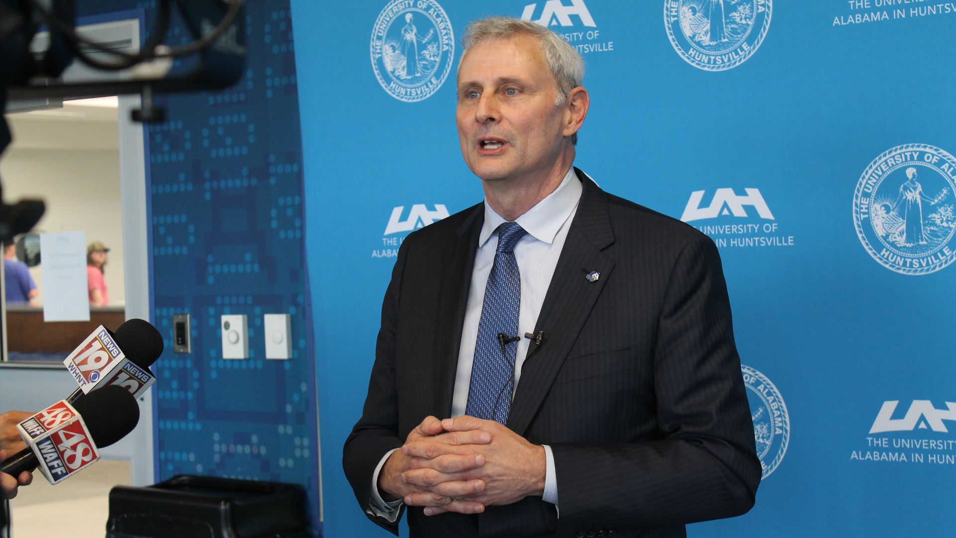 Man in a dark suit and blue tie speaks at a press conference, standing before a blue backdrop with the University of Alabama in Huntsville logos; microphones visible in the foreground.