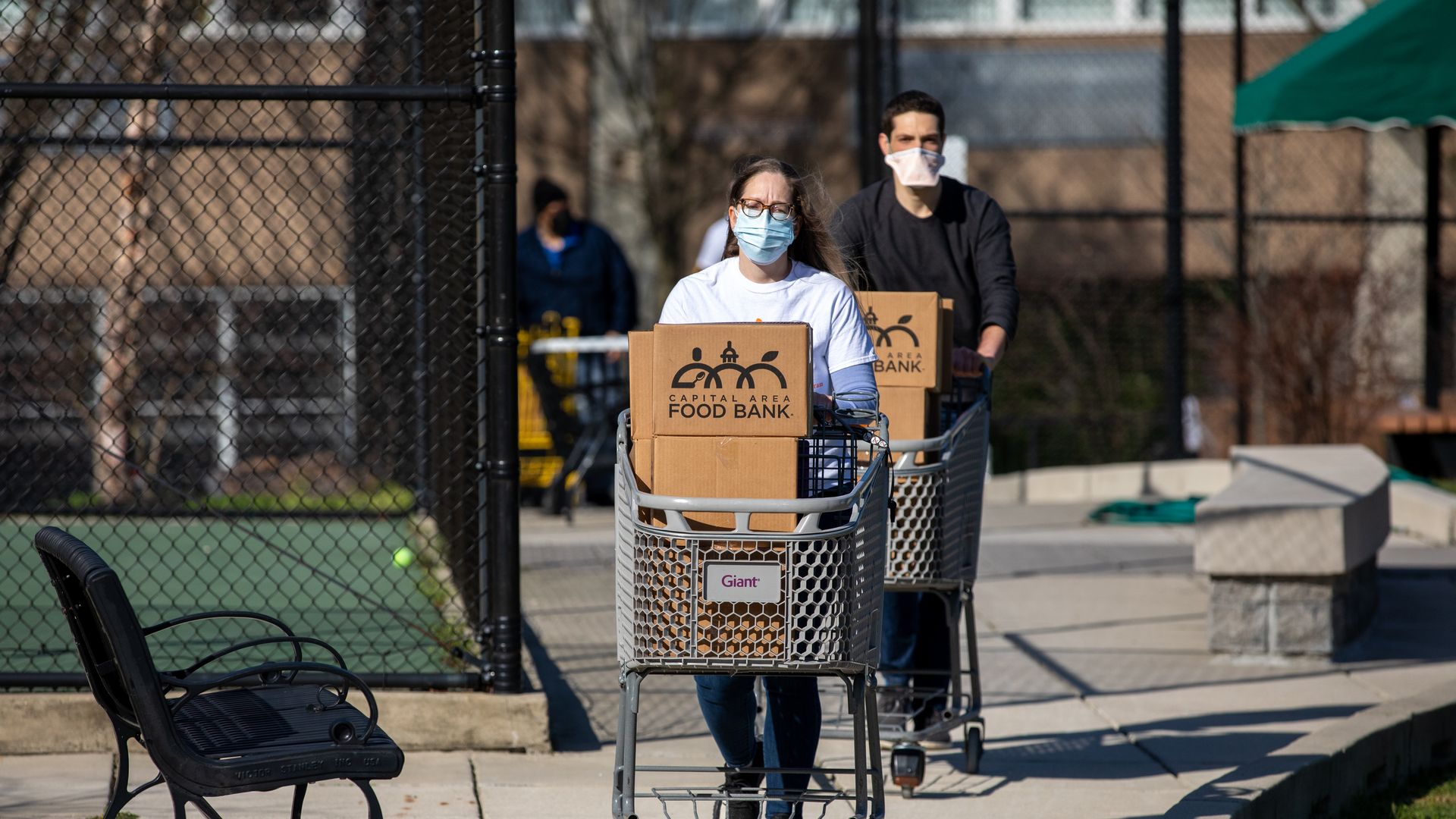 Two people, one in front of the other, push shopping carts piled with brown boxes that say FOOD BANK on them.