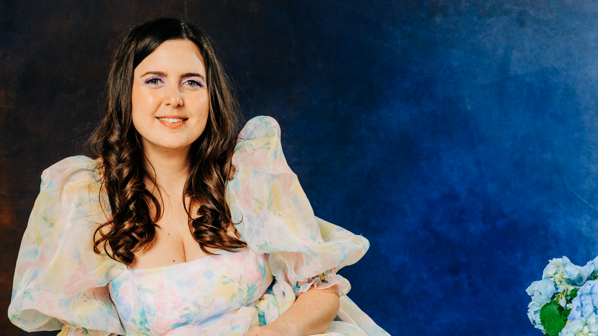 Maddy Brannon poses in a colorful dress with puffy sleeves, in front of a blue background.