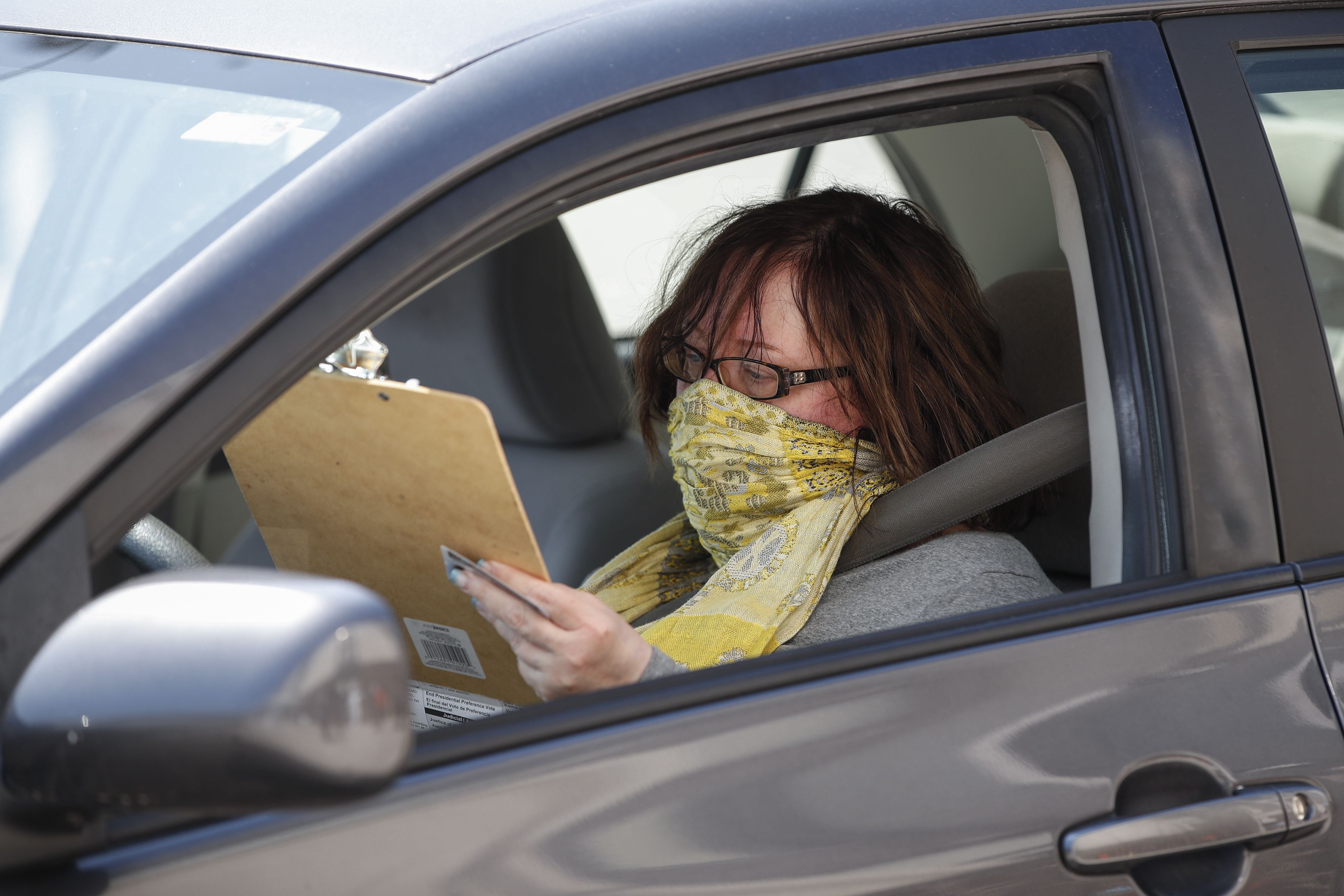 In this image, a woman sits in her car while wearing a bandana around her face and filling out a clipboard