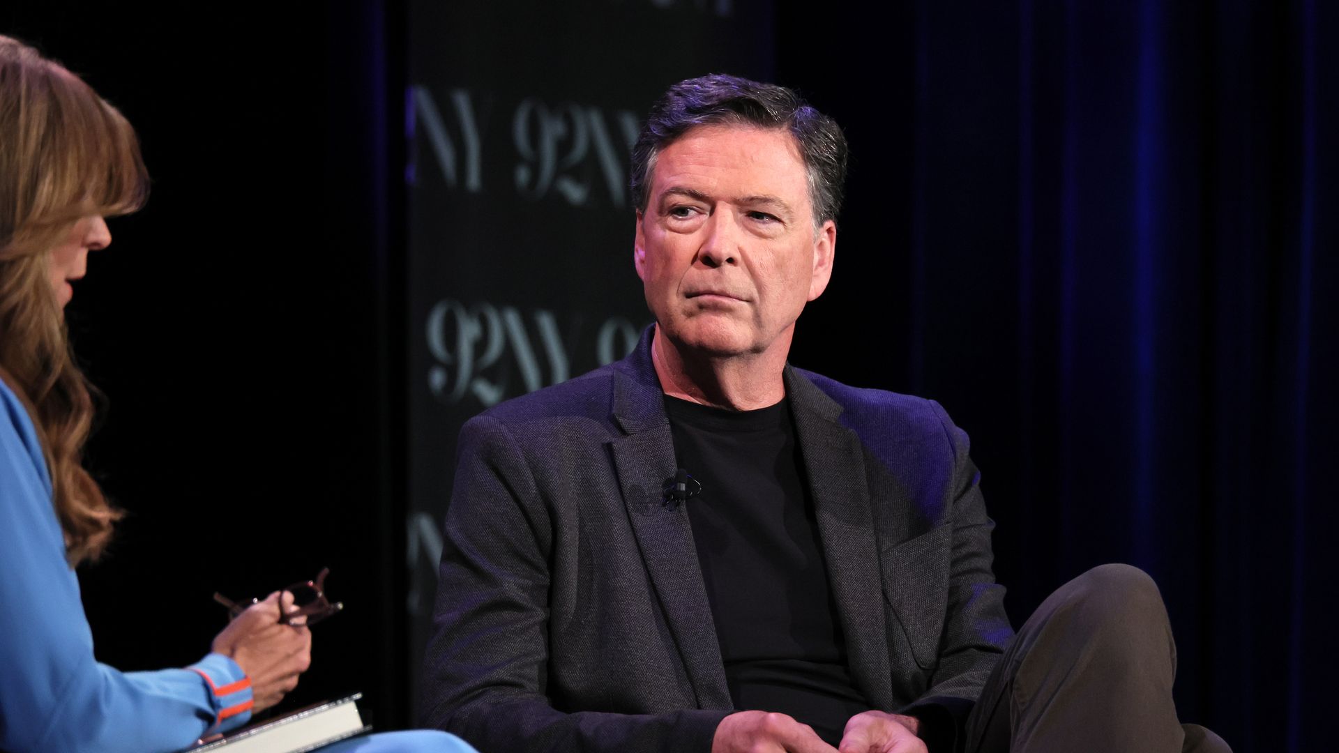 James Comey, wearing all black, sits as he is interviewed on a stage by a woman in blue.