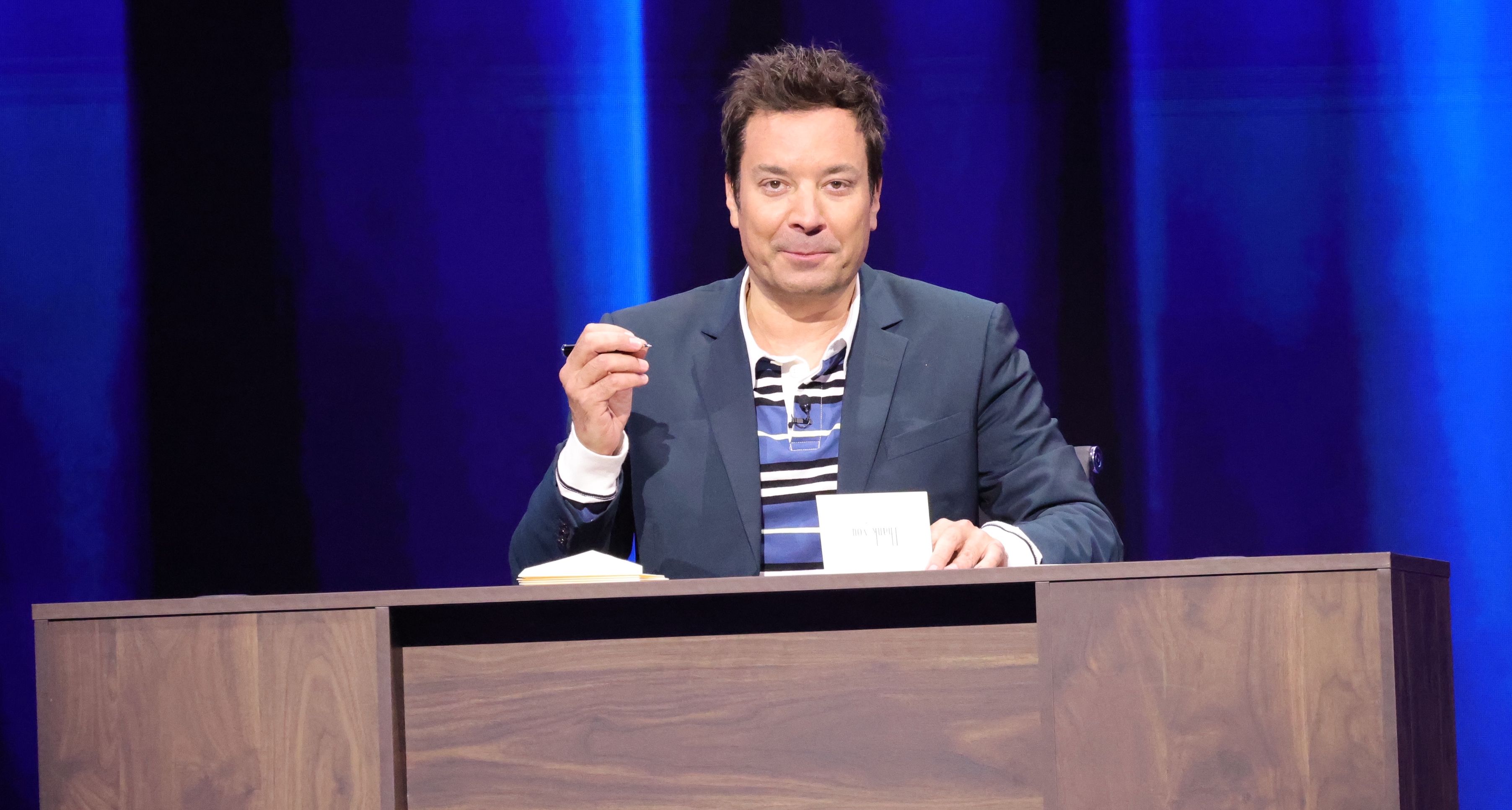 Jimmy Fallon in blazer sits at a wooden desk on stage, holding a pen with blue lights behind him, appearing mid-show.