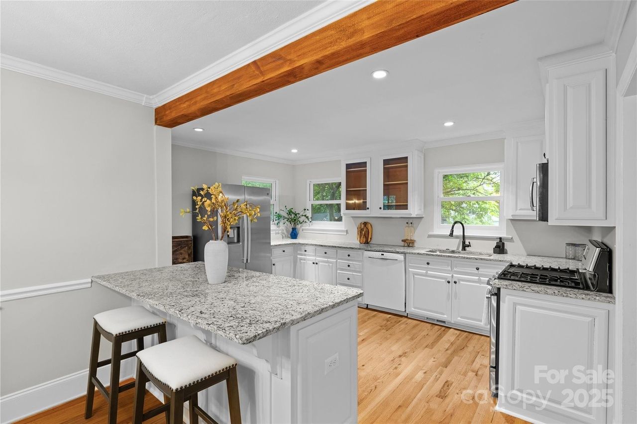 Bright kitchen with white cabinets, granite countertops, a wooden floor, stainless steel refrigerator, stove, and kitchen island with two stools and a white vase with yellow flowers.