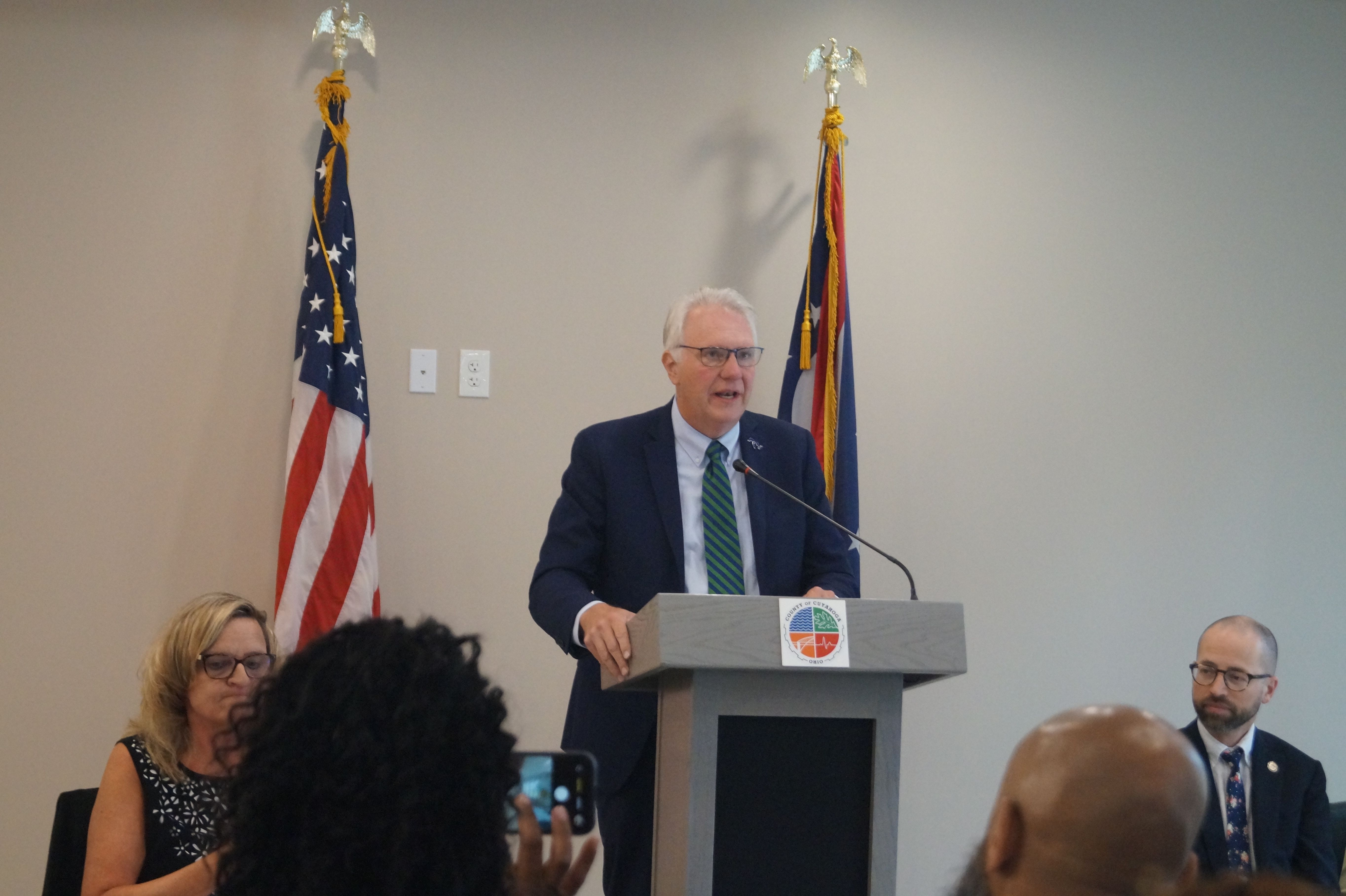 A man in a dark suit and green striped tie speaks at a podium, flanked by two flags (U.S. flag on the left). Audience sits, with a person in the foreground recording on a phone.