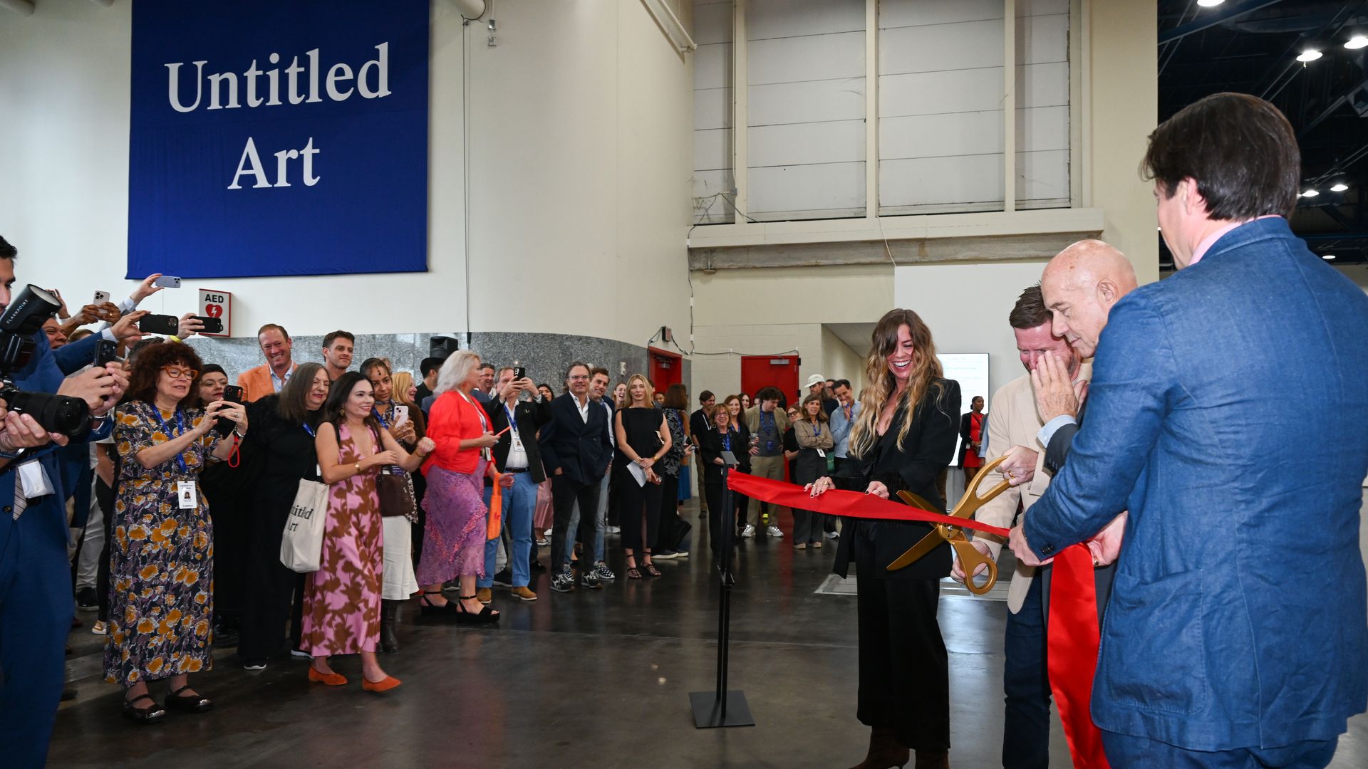Inside a gallery, a ribbon-cutting ceremony as a crowd watches. A large blue sign says 'Untitled Art'; a woman and others hold phones while a man in a blue suit cuts a red ribbon.